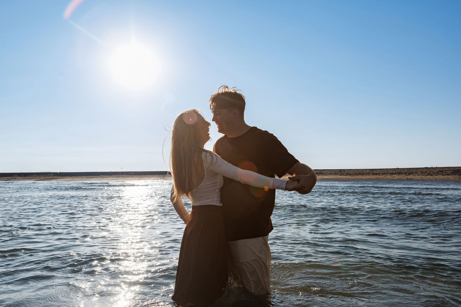 A silhouetted couple embraces at the water's edge with sun glinting off the ocean surface on a bright day.