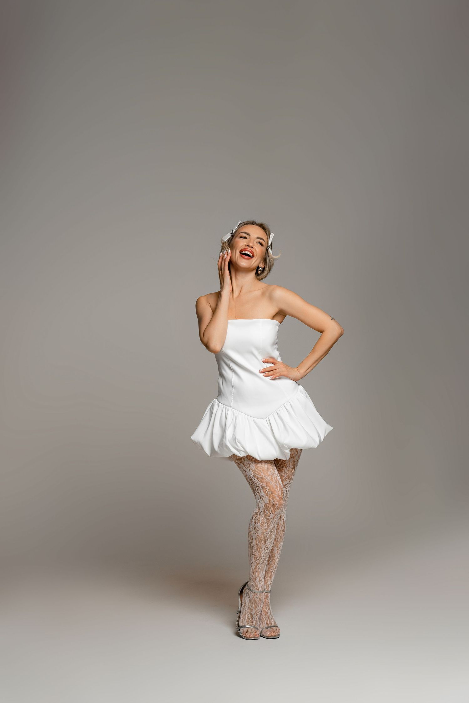 Woman in a short white strapless dress and patterned stockings poses playfully on a gray studio background.