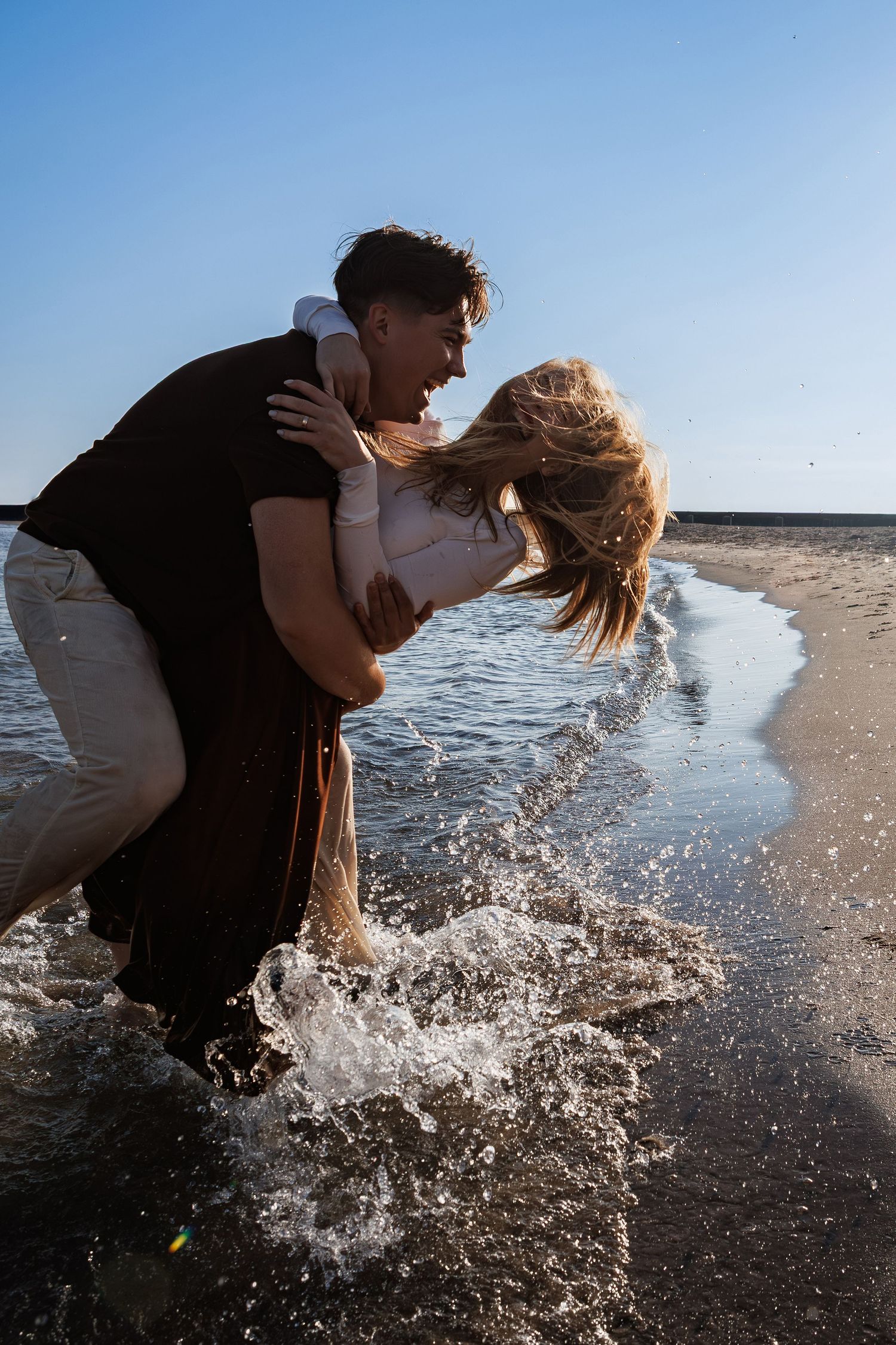 A couple embraces playfully at the beach while splashing through shallow ocean waves at sunset.