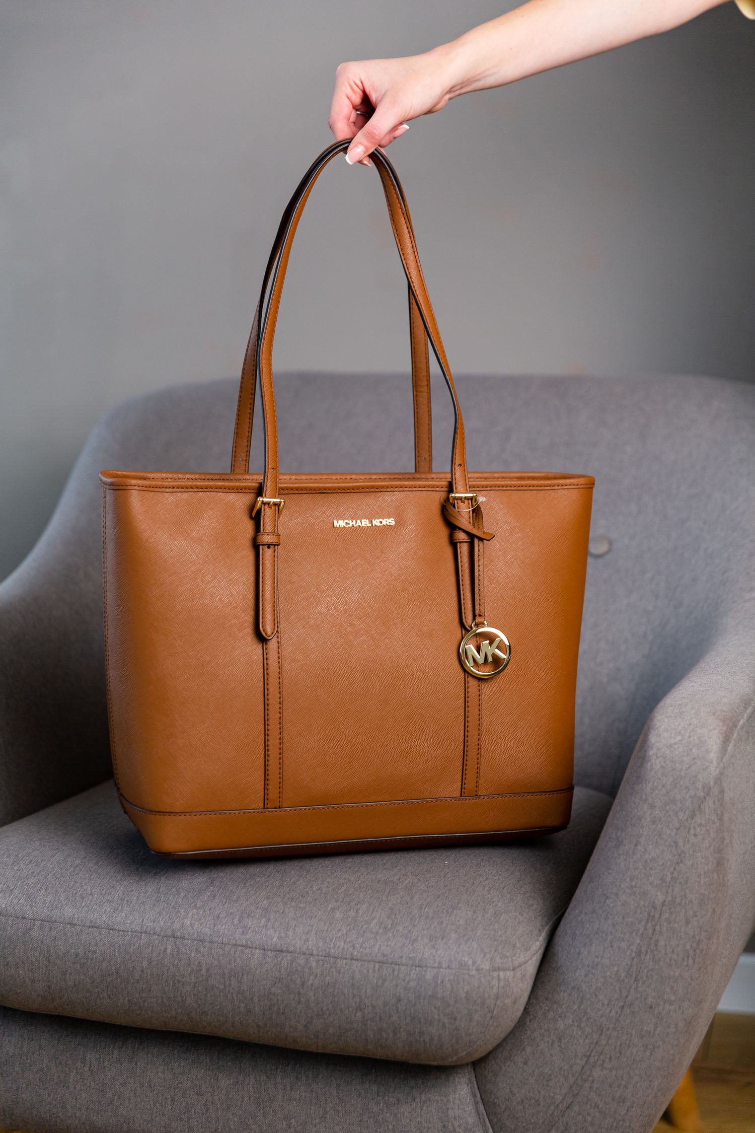Cognac brown Michael Kors leather tote bag with signature charm displayed on gray chair.