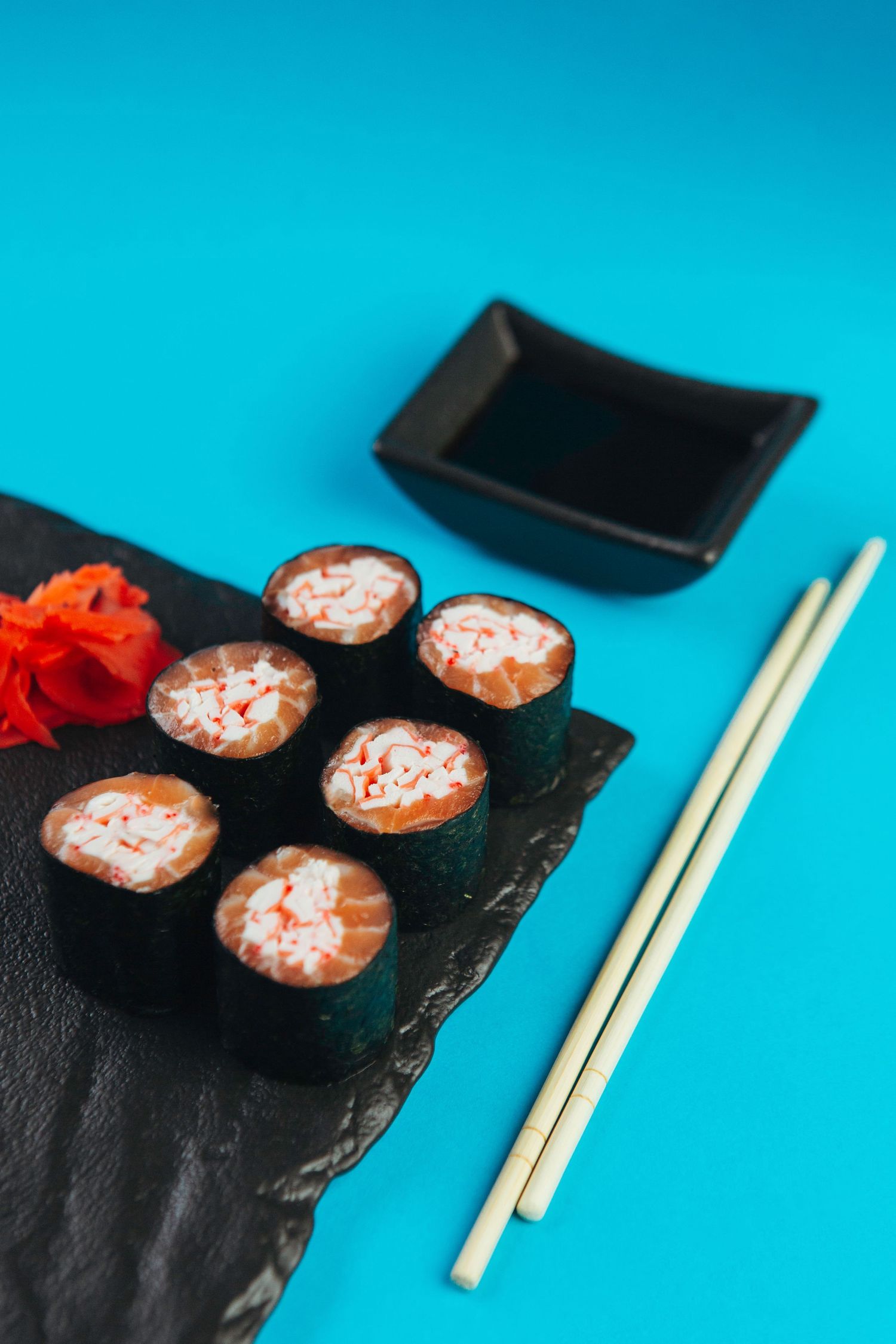Sushi rolls plated on black slate with chopsticks against bright blue background.