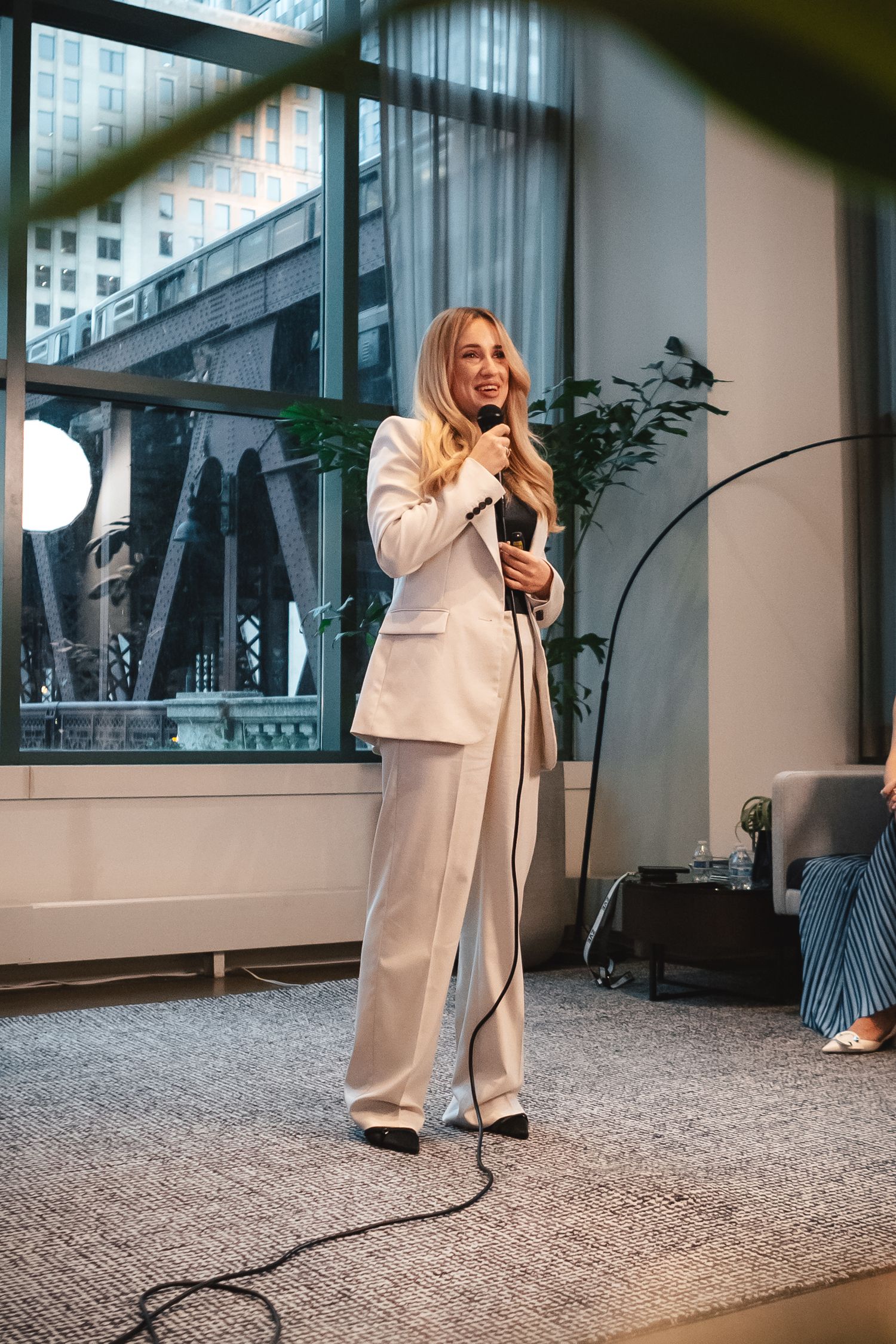 A person in a cream colored business suit speaks into a microphone during a presentation in a modern office space.