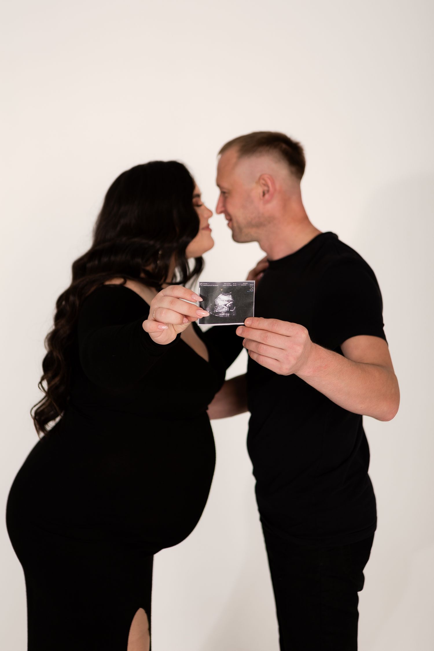 Couple sharing intimate moment while holding ultrasound photo during maternity shoot.
