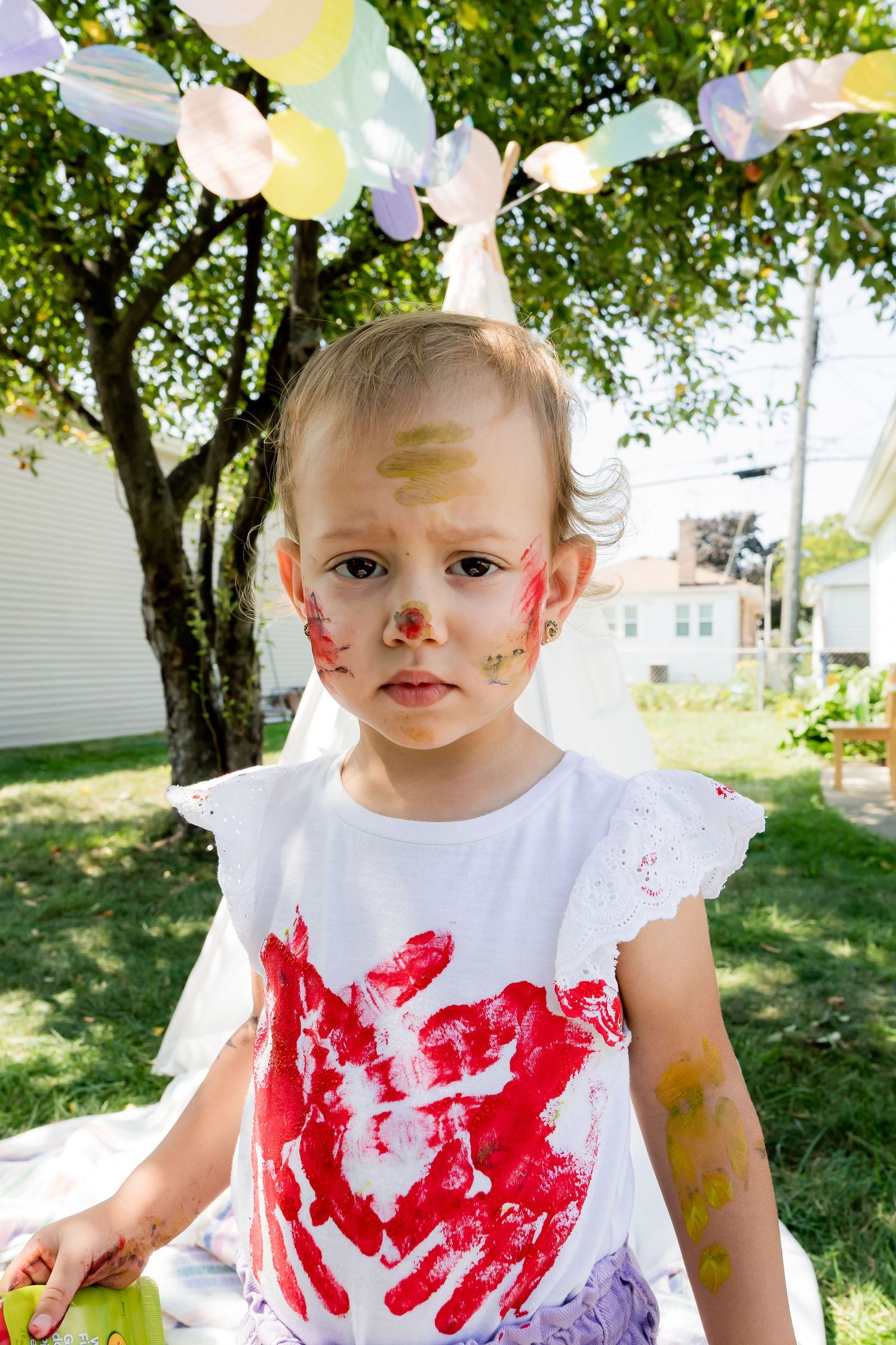 Small child in white shirt covered in colorful paint stains under party decorations.