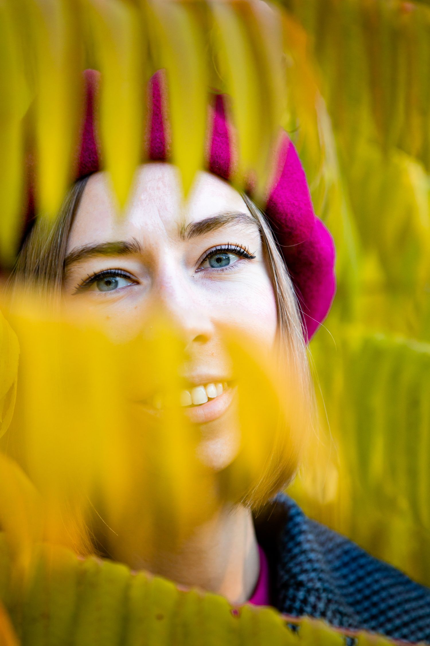 Creative portrait through yellow autumn leaves showing vibrant magenta beret and natural lighting.