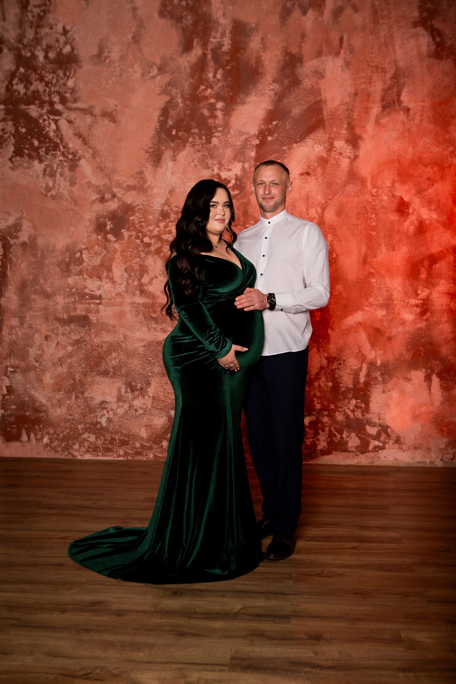 Maternity portrait with woman in emerald velvet gown against rustic backdrop.