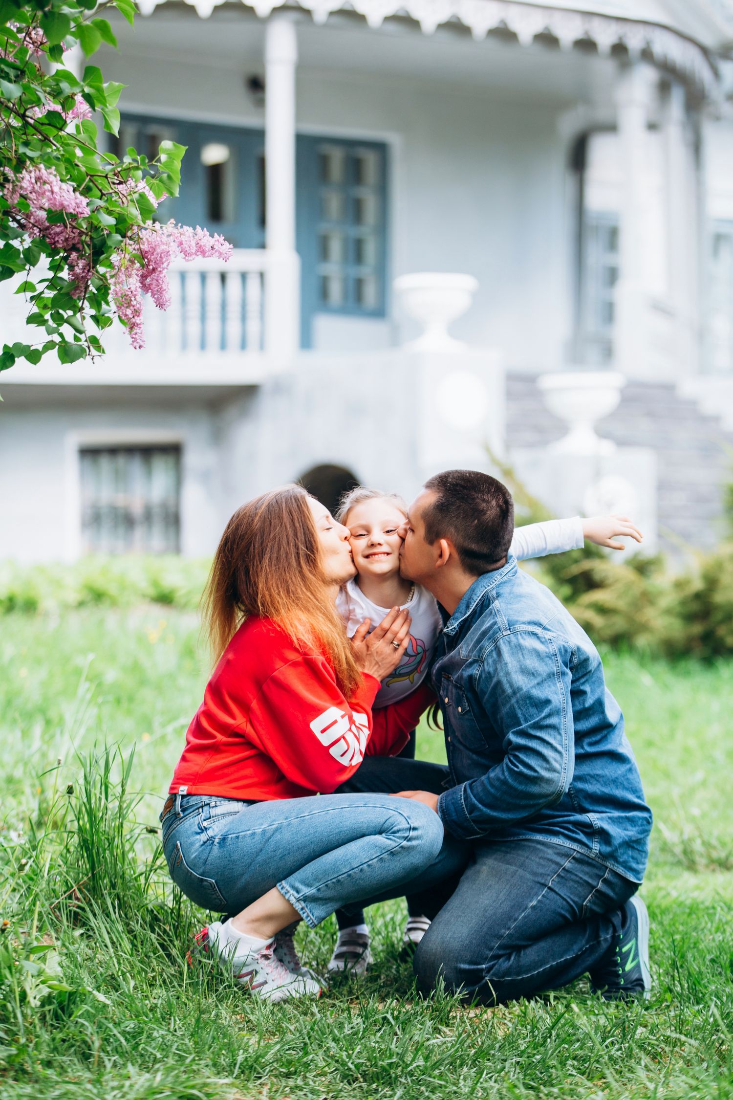 A sweet family moment shared on the lawn with blooming lilac bushes and a white house in the background.