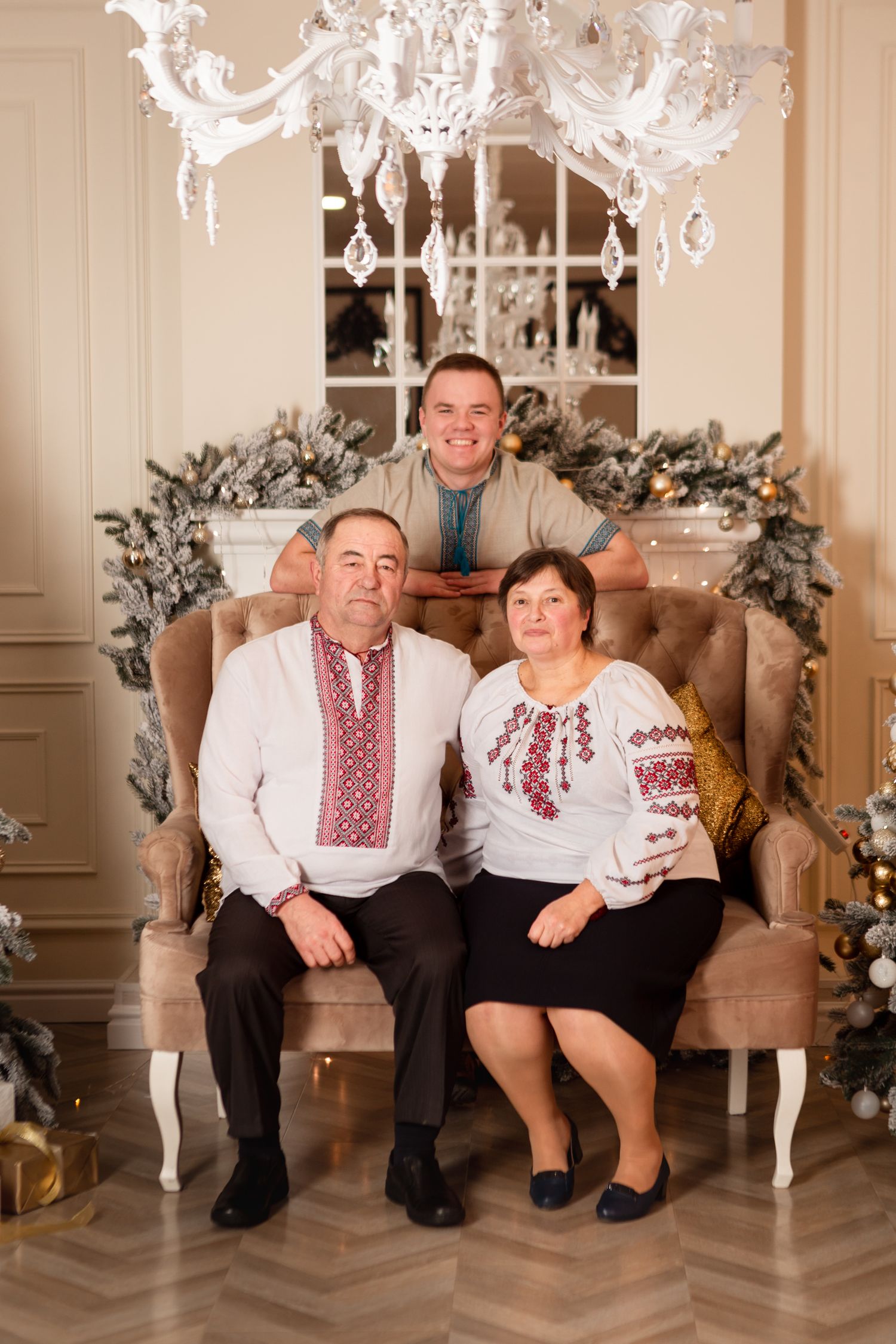 Family portrait in traditional embroidered attire seated on a beige chair under an elegant crystal chandelier.