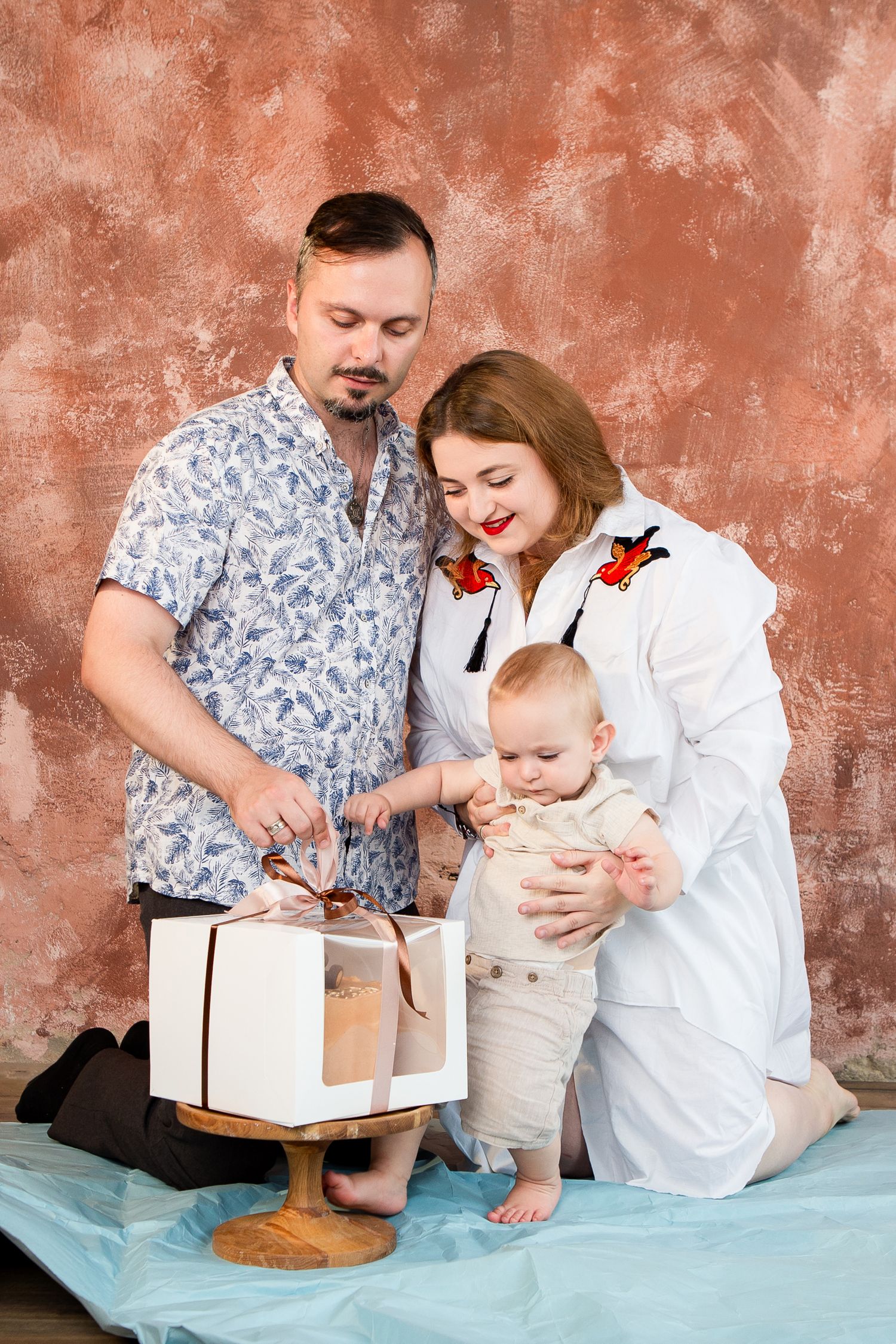 Family celebrating first birthday with a cake on blue backdrop against textured coral wall.