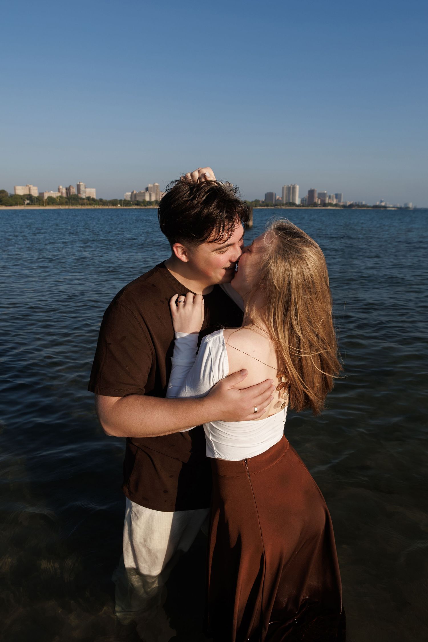 Romantic couple embraces while standing in shallow ocean waters with city skyline visible in background.