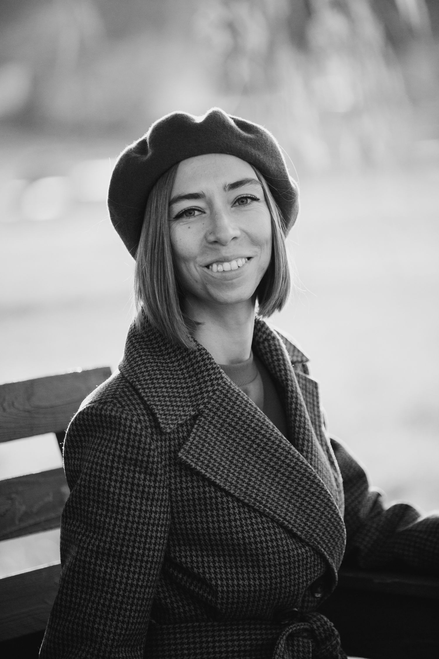 Person wearing black beret and plaid coat sitting on bench in black and white portrait.