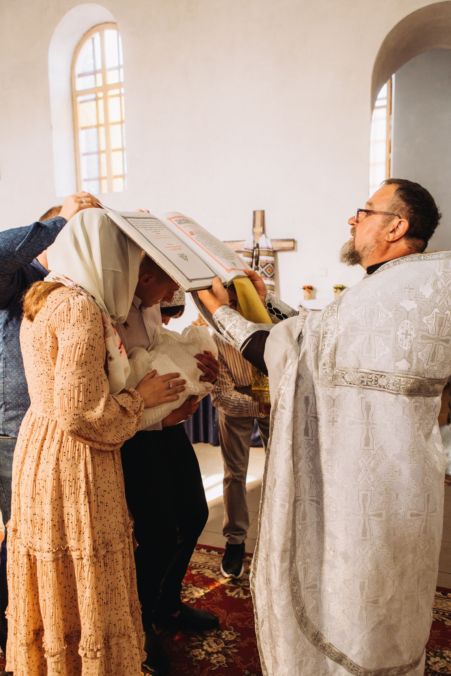 An Orthodox priest in white robes performs a religious ceremony in a church with arched windows.