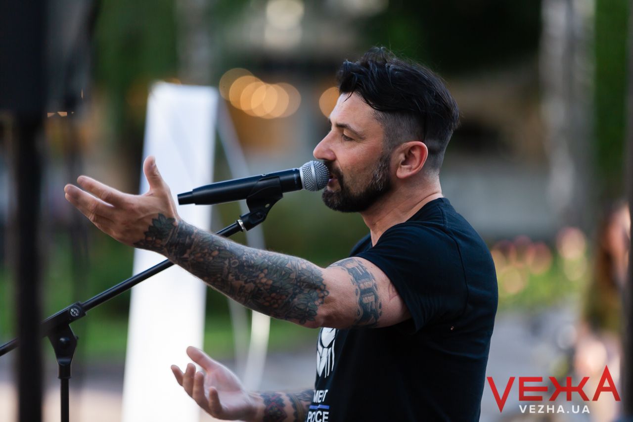 A performer singing into a microphone on stage with tattooed arms gesturing during an outdoor performance.