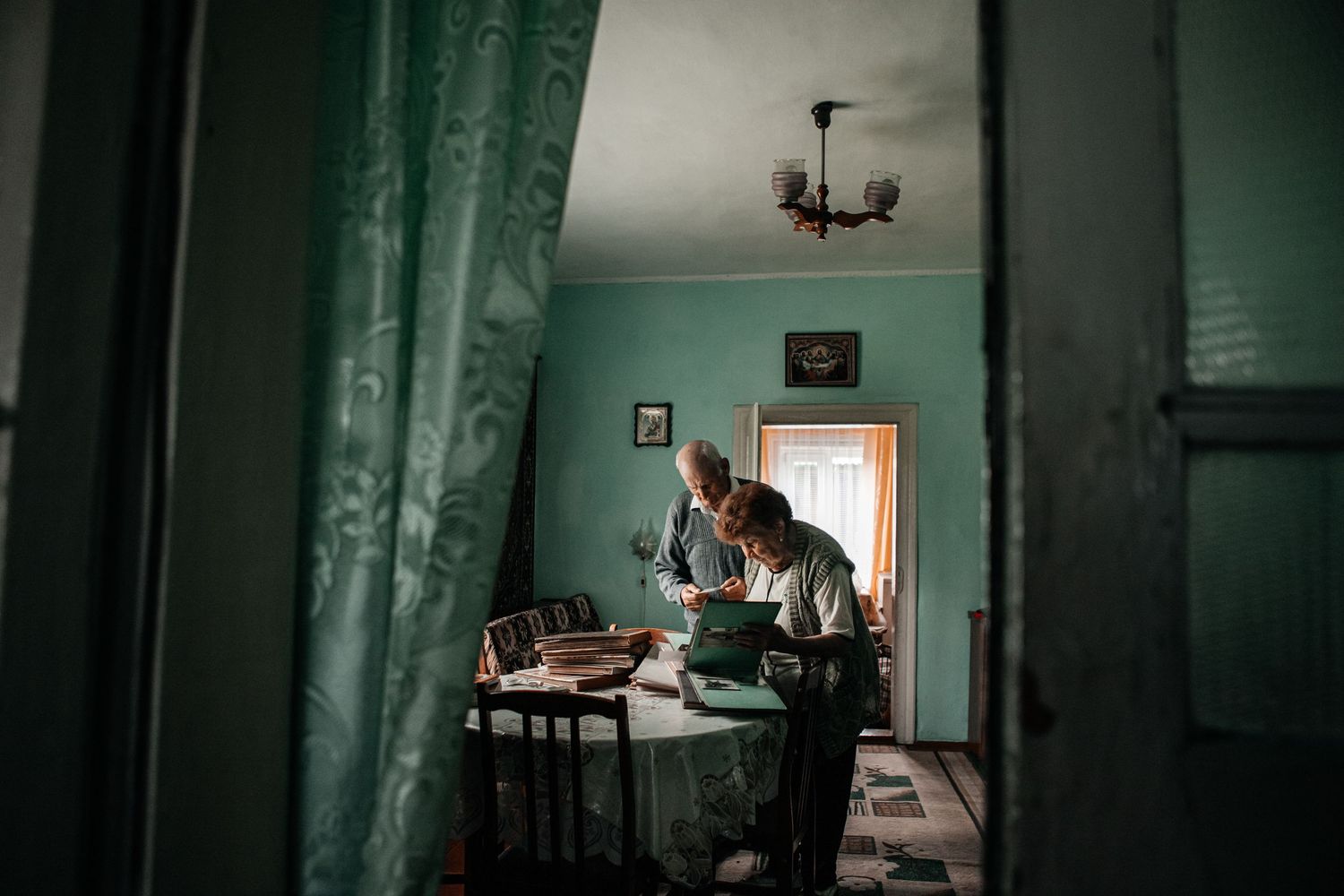 Interior view of a room with green curtains and someone sitting at a table near a window.