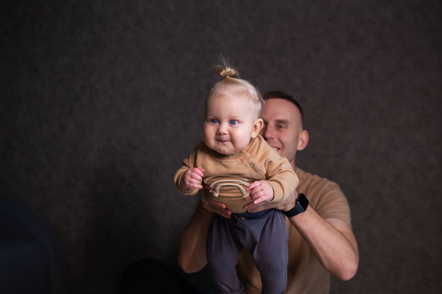 A baby wearing a beige sweater smiles while being held against a dark background.