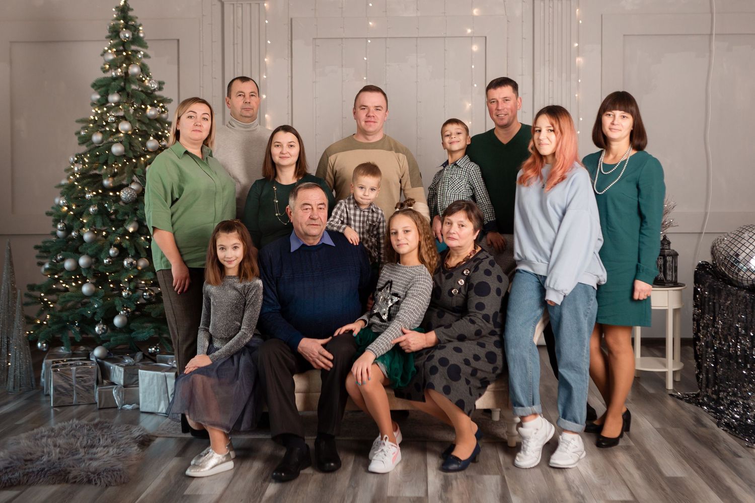 Large family group portrait in front of decorated Christmas tree in grey and white studio setting.
