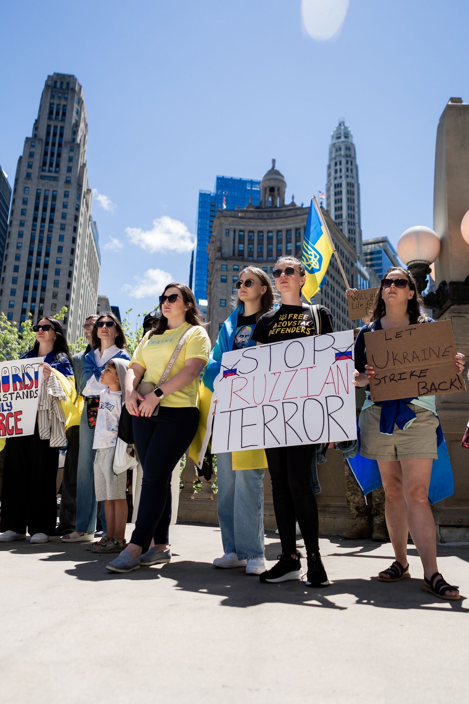 Demonstrators hold protest signs with Ukrainian flags against a backdrop of Chicago skyscrapers.