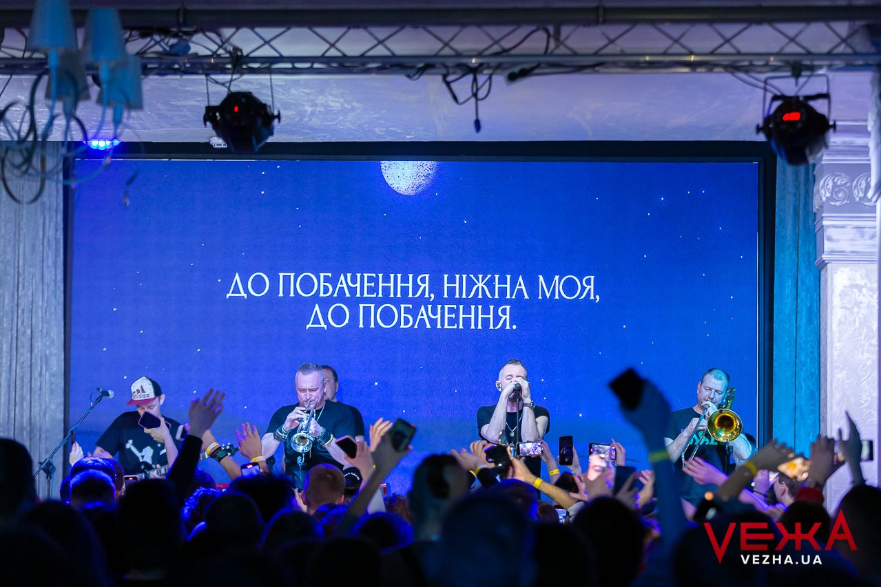 Band performs on stage with blue lighting and text projection behind them.
