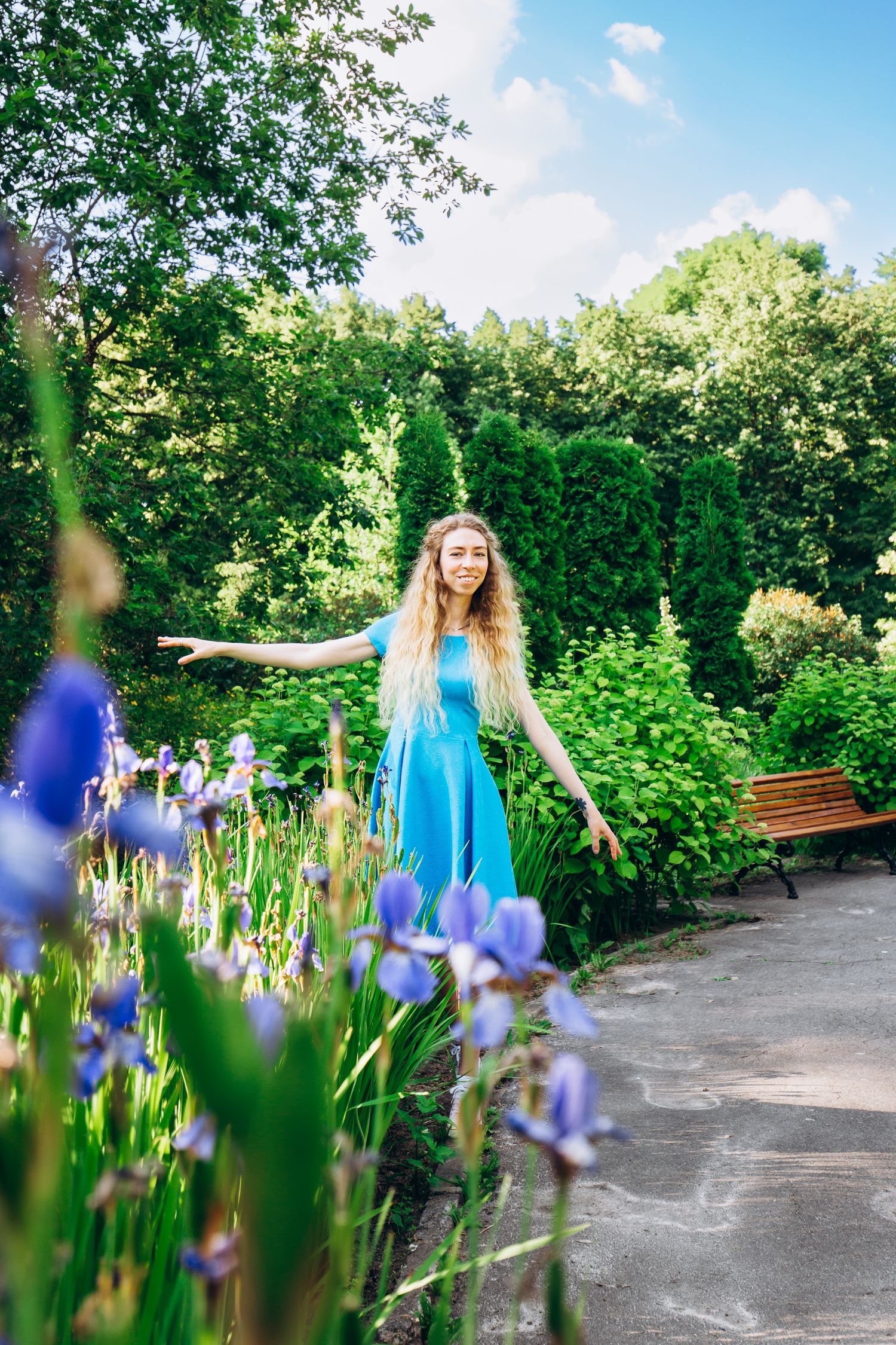 Person in blue dress dancing among purple iris flowers in sunny garden pathway.