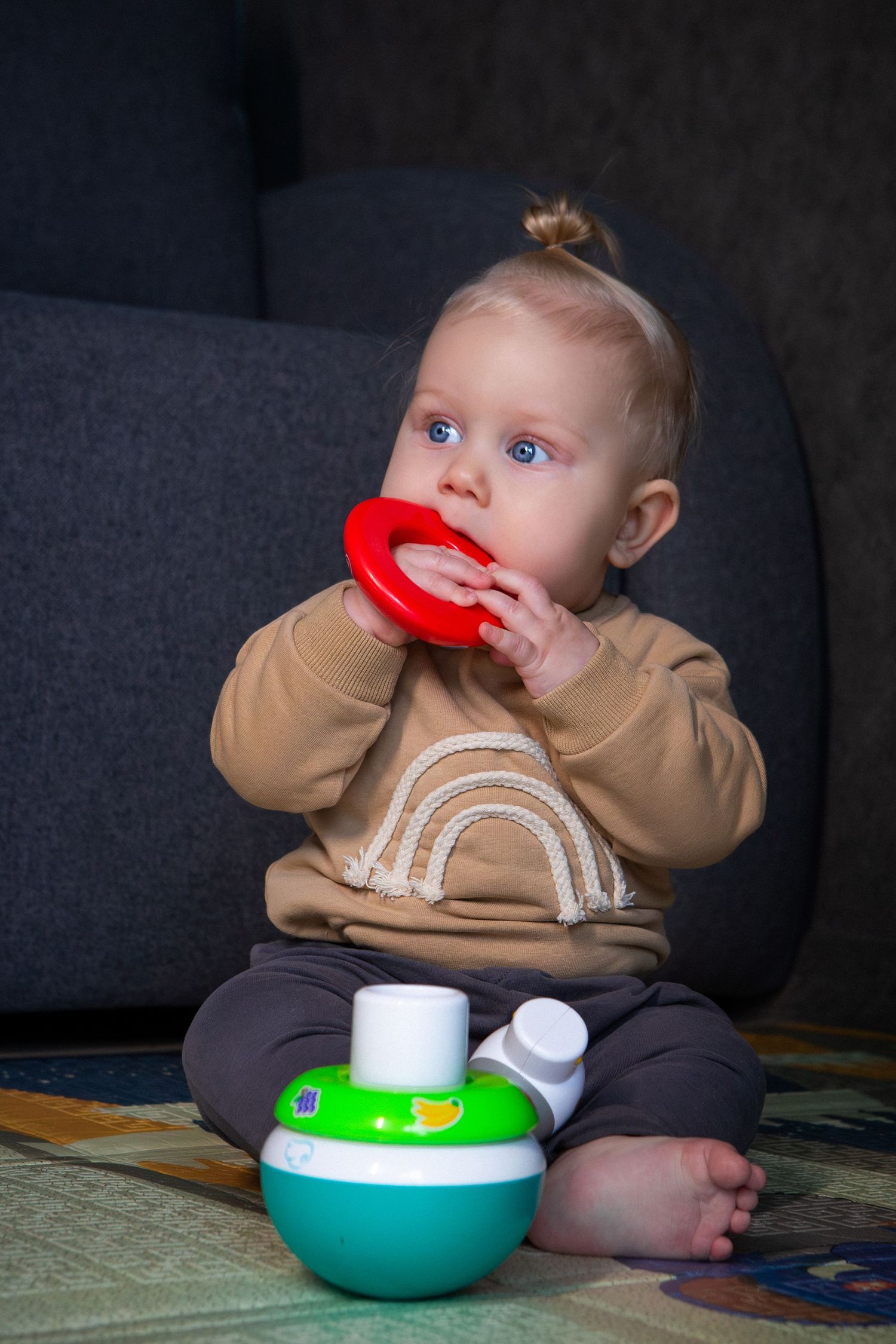 A baby in a tan sweatshirt plays with a colorful interactive toy on a dark couch.