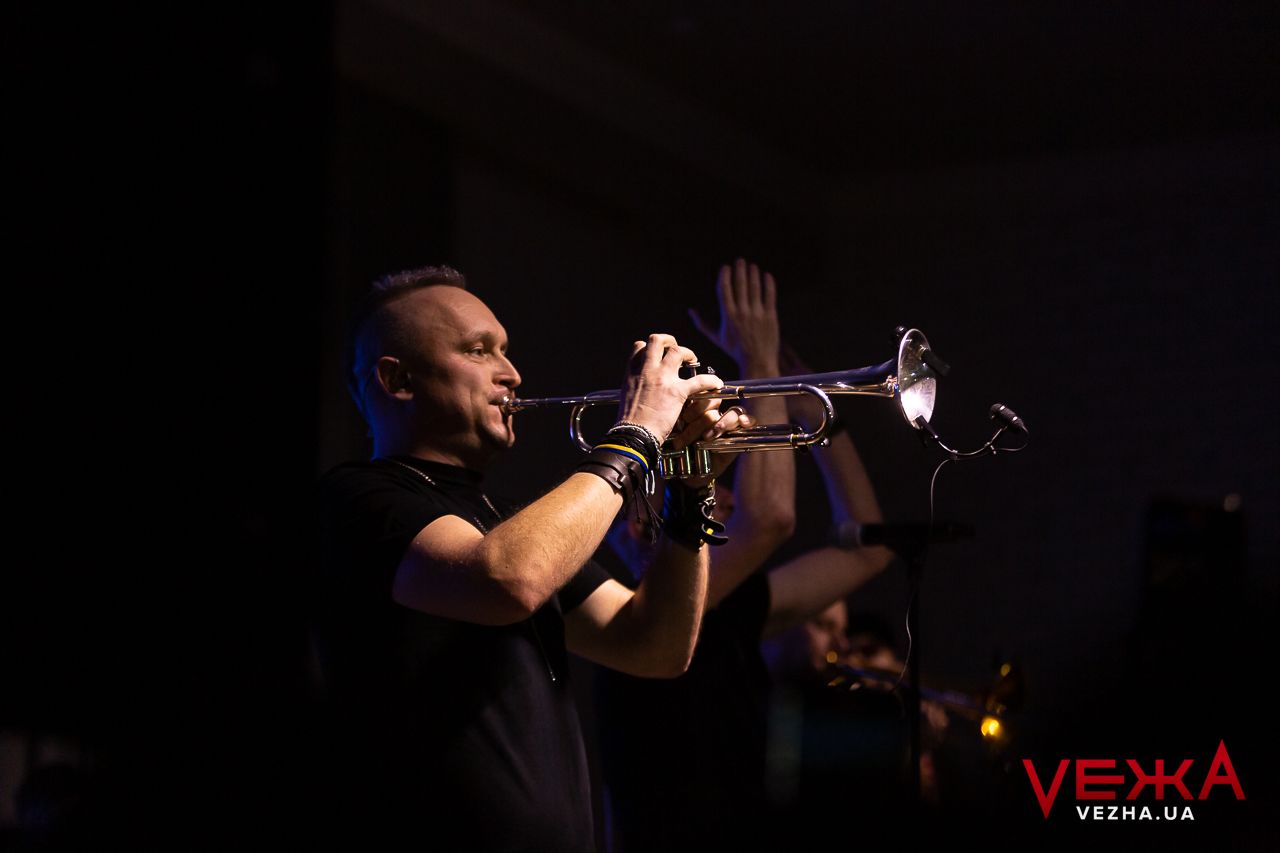 A musician plays trumpet on a dark stage with dramatic lighting.