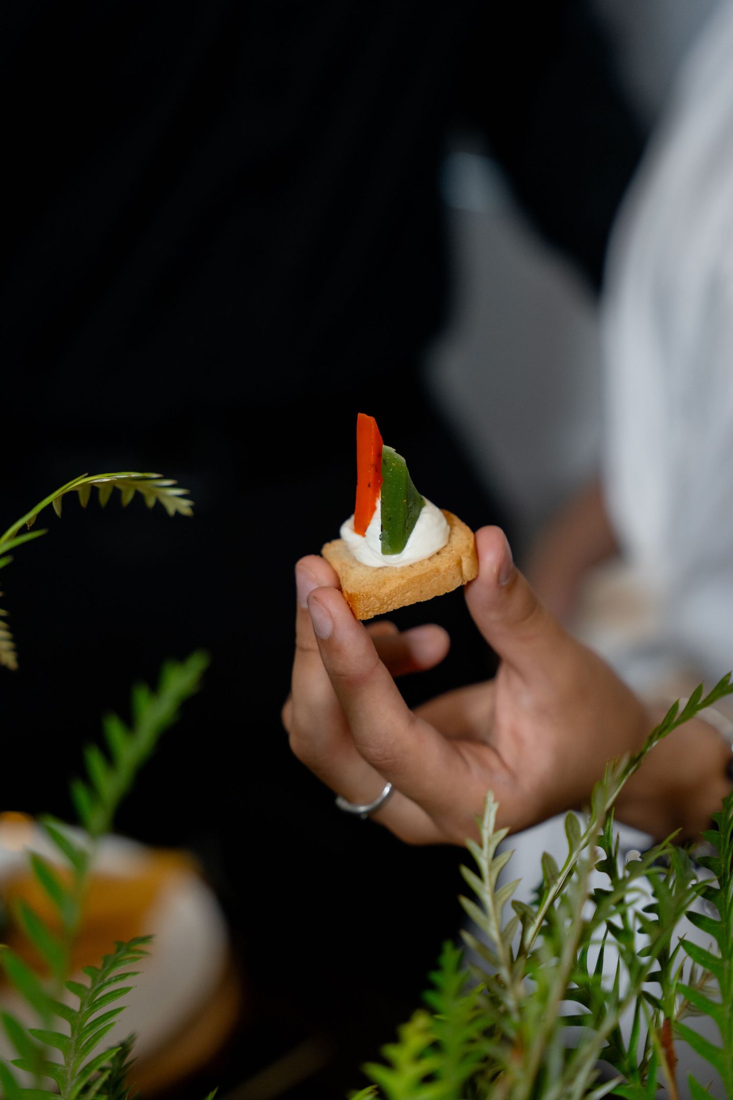Close-up of hand holding appetizer with cream and red garnish among fern leaves.
