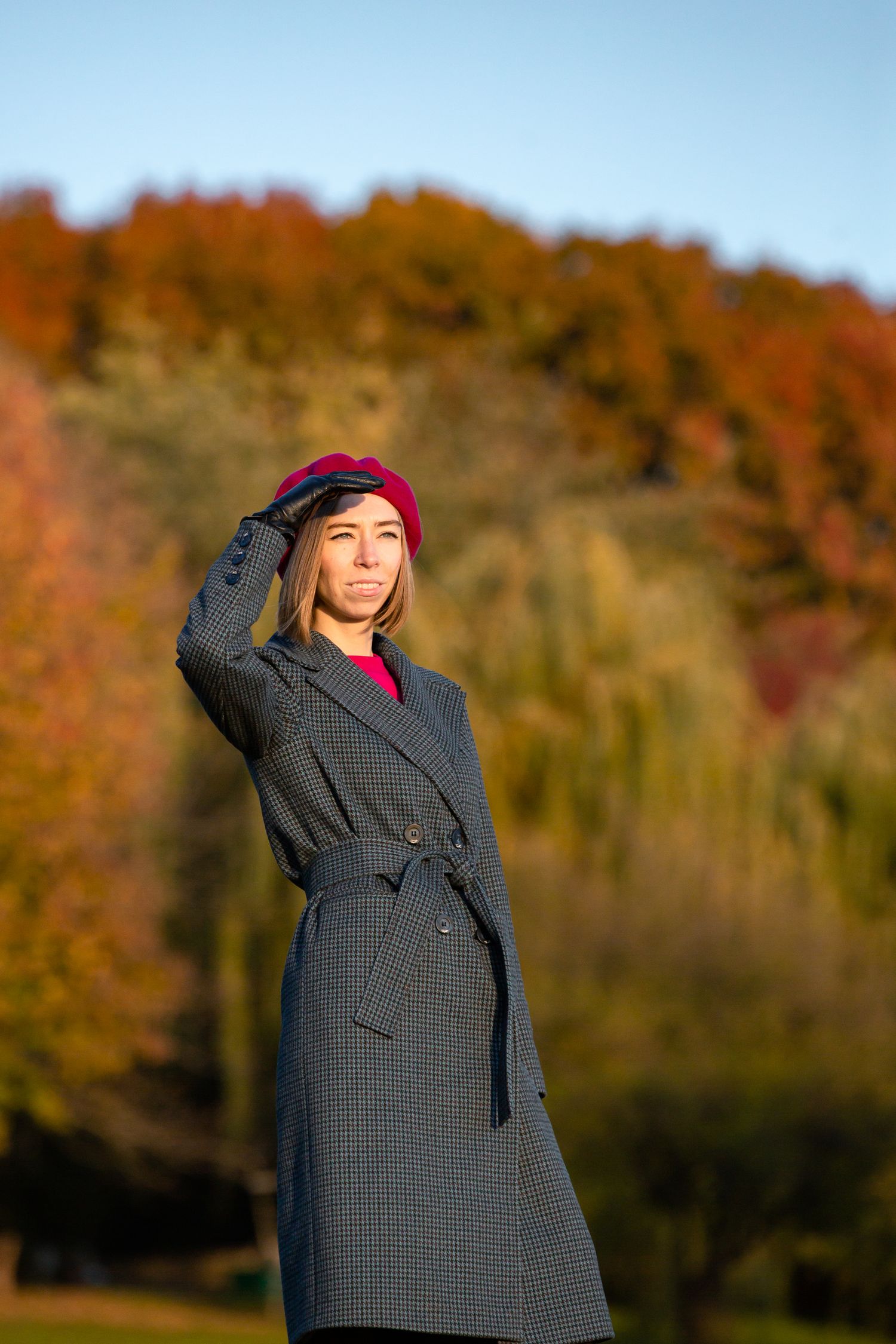 Person in long coat and burgundy beret poses against autumn landscape.