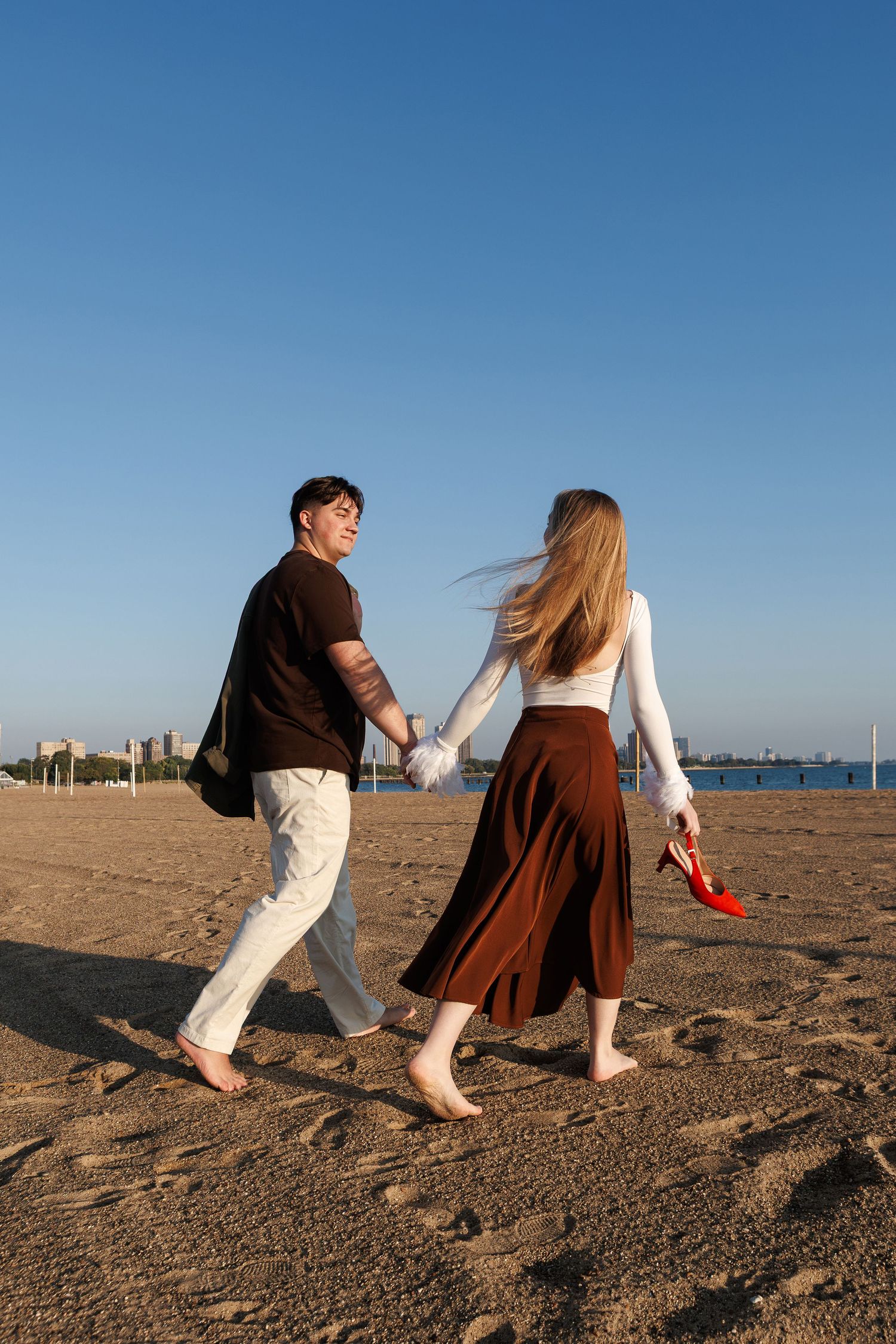 Couple walks barefoot along sandy beach at sunset holding red shoes.