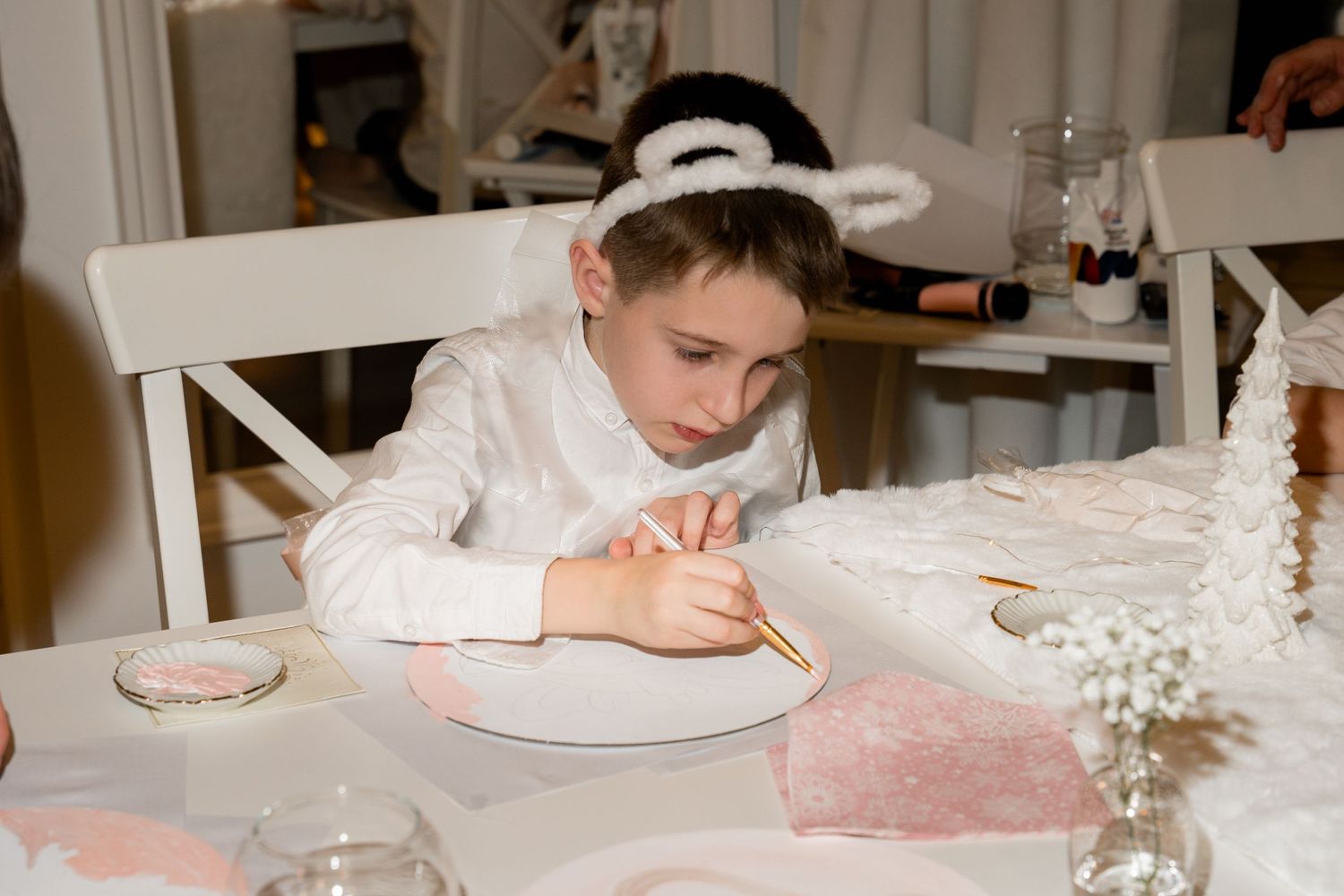 Child in white writing at table with pink and white party decorations.