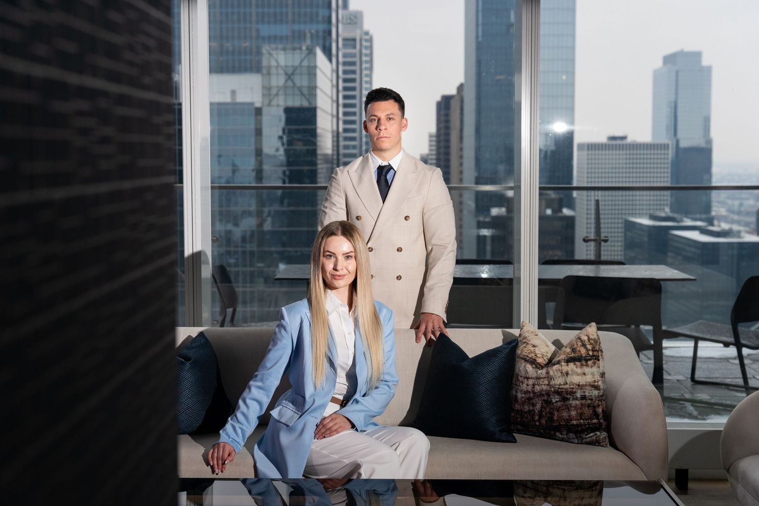 Professional business pair in contrasting suits pose on modern office couch with city skyline backdrop.