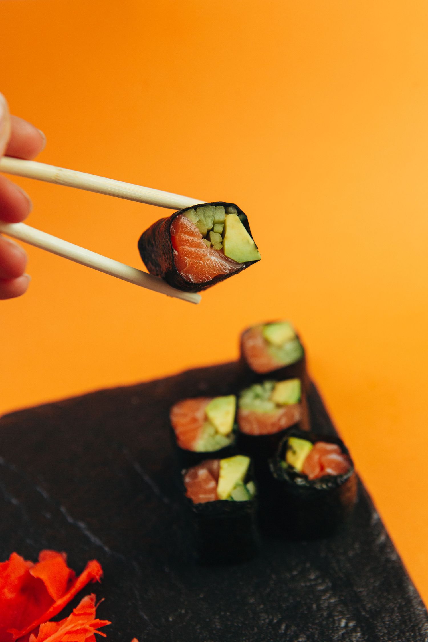 Chopsticks holding salmon and avocado sushi roll against vibrant orange background.