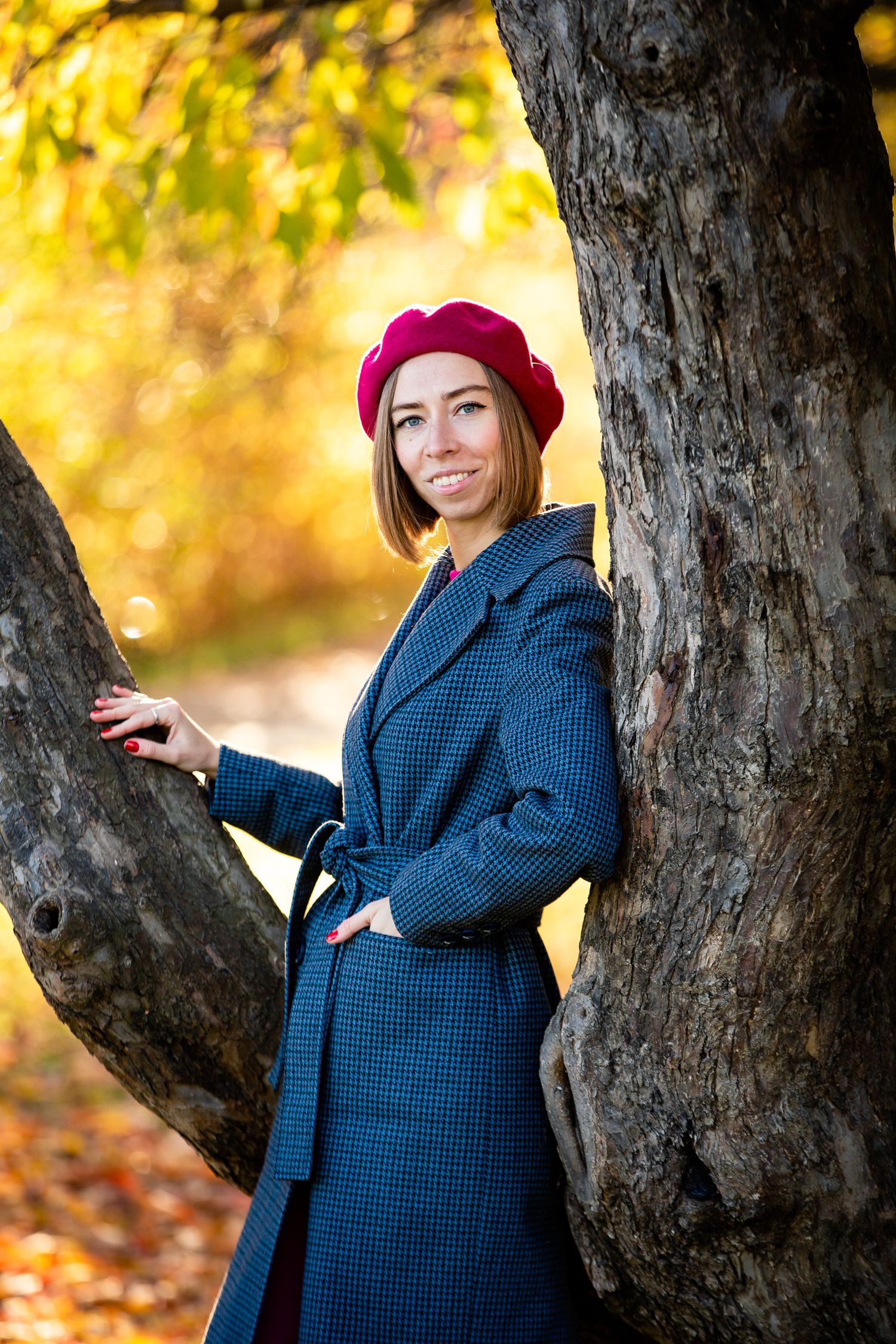 Person in plaid coat and red beret posing against tree trunk with autumn foliage.