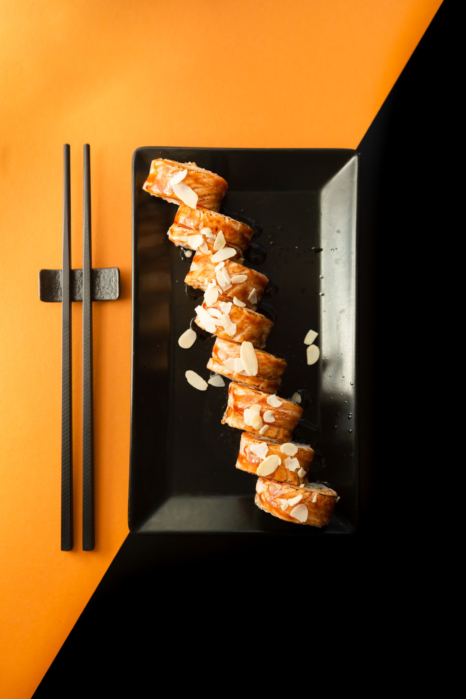 Diagonal arrangement of sushi rolls on a black rectangular plate with chopsticks against an orange and black background.