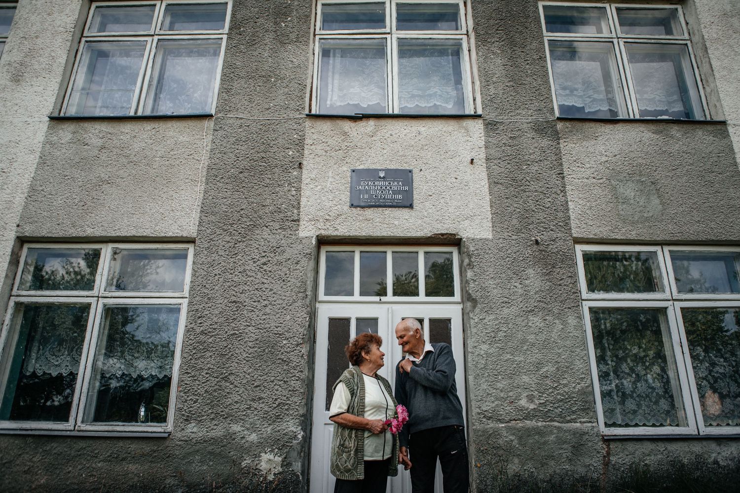 Two people standing in conversation at a window of an old concrete building.
