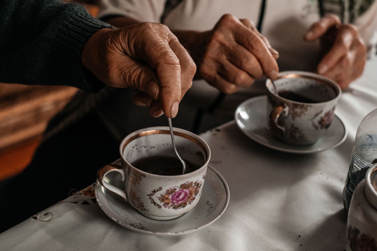 Close-up of hands stirring tea in decorative porcelain cups on white tablecloth.