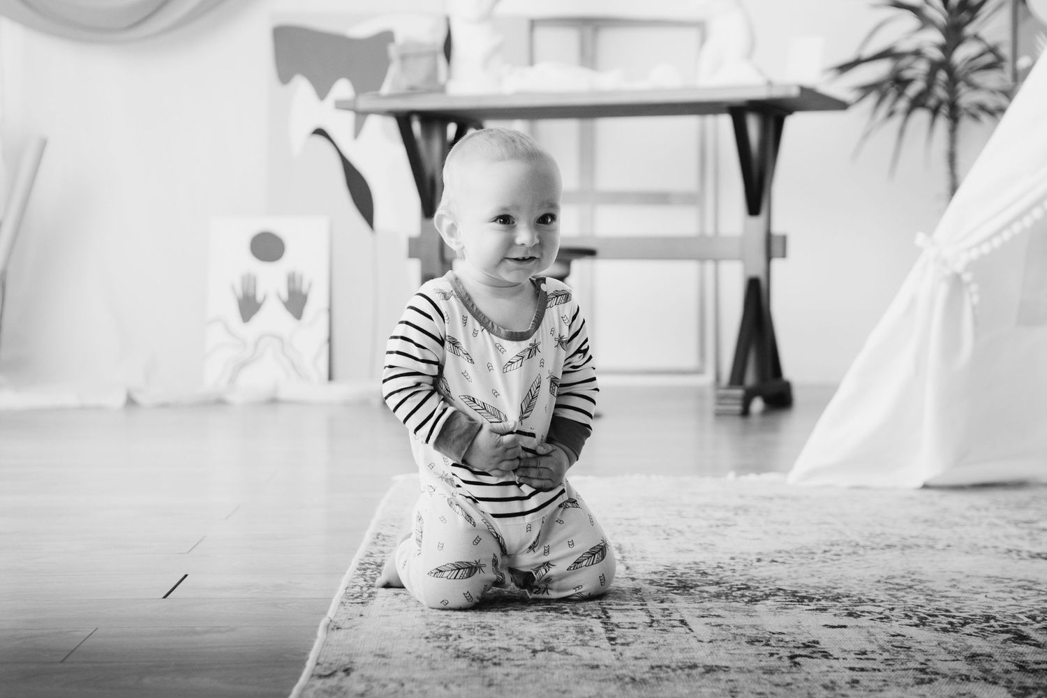 Black and white photo of smiling baby in striped shirt crawling on floor.