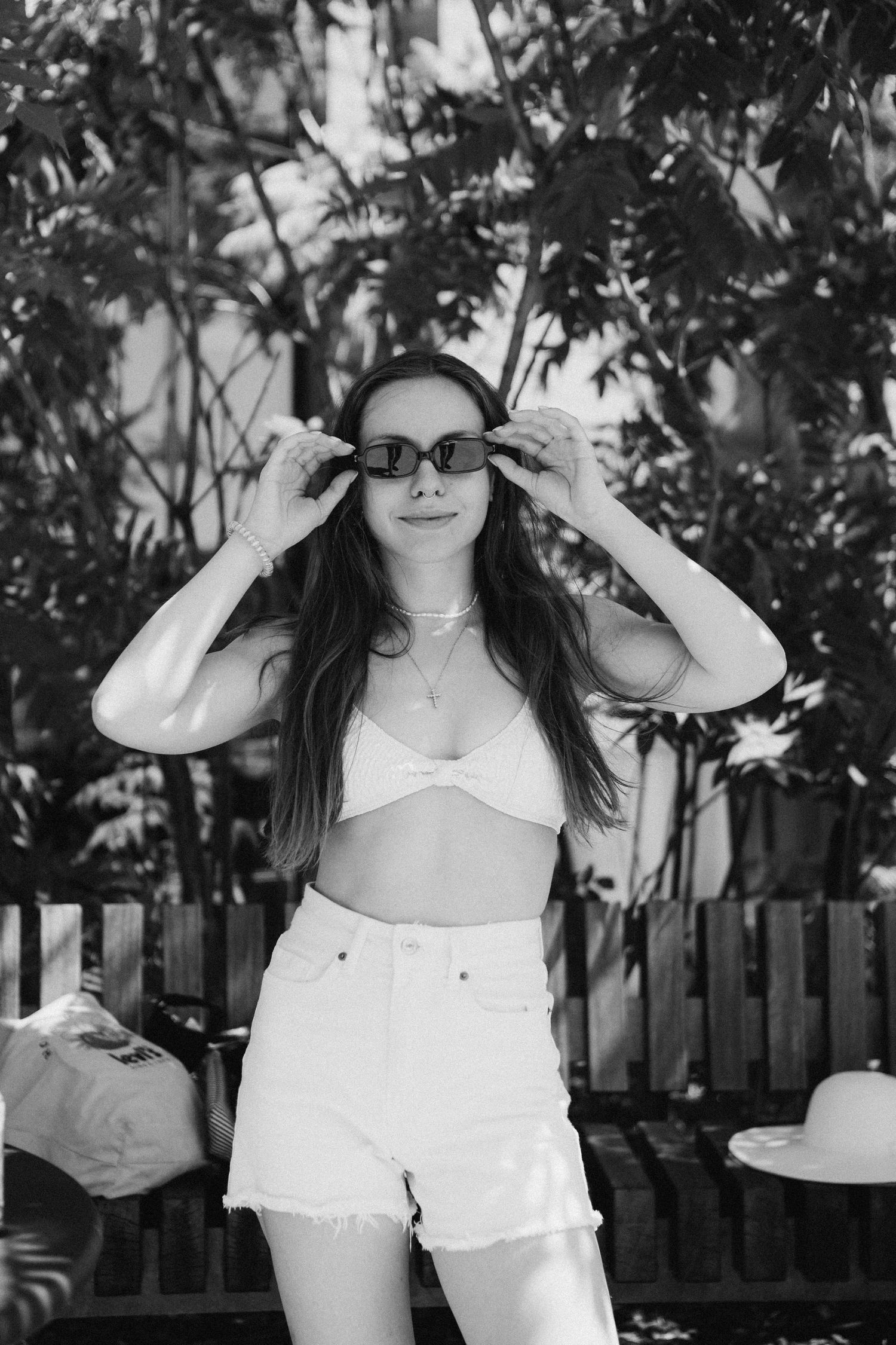 Black and white photo of someone wearing sunglasses, a white bikini top and high waisted shorts in a garden setting.