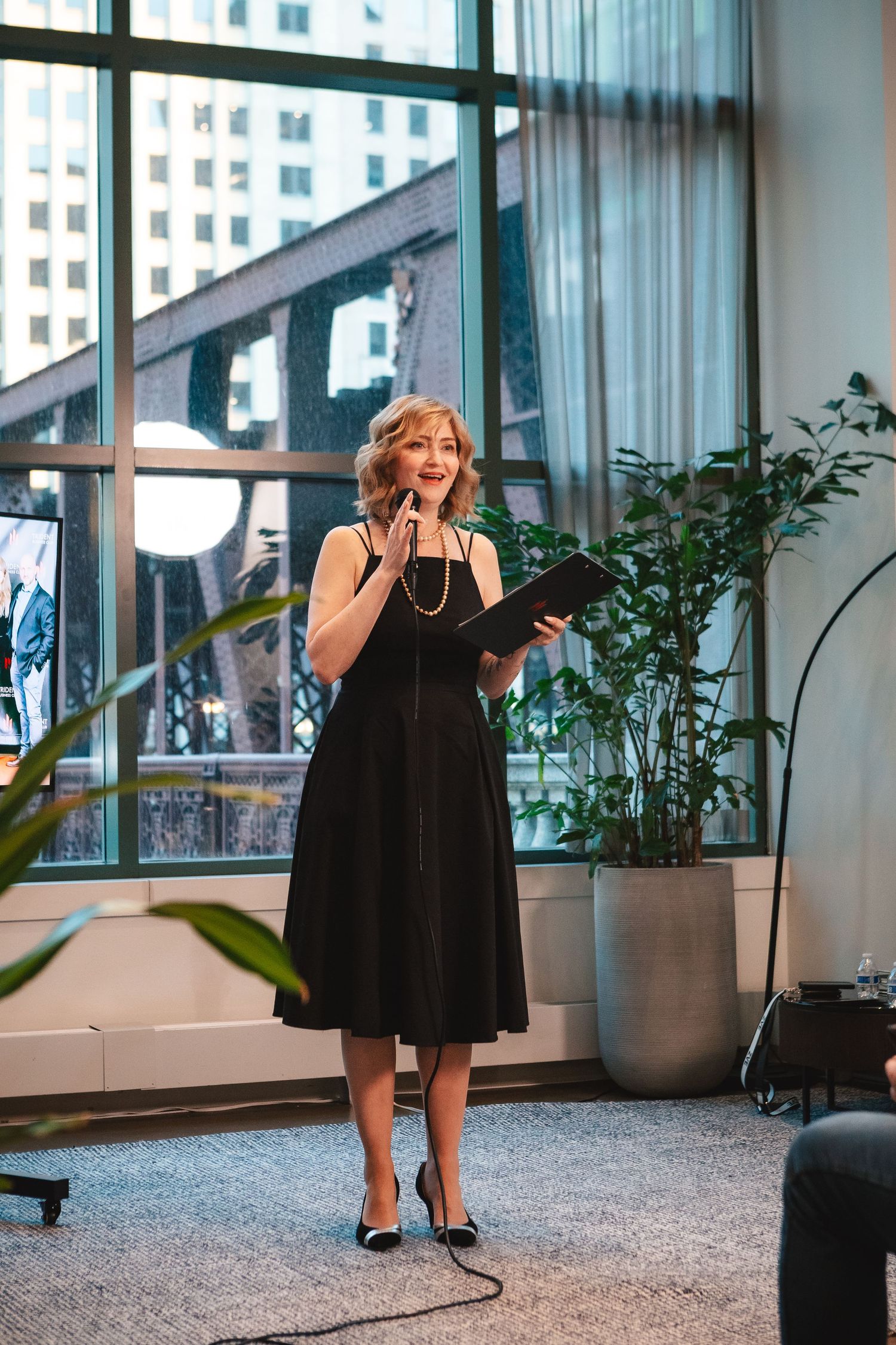 Speaker in a black dress presenting to an audience in a modern office space with city views.