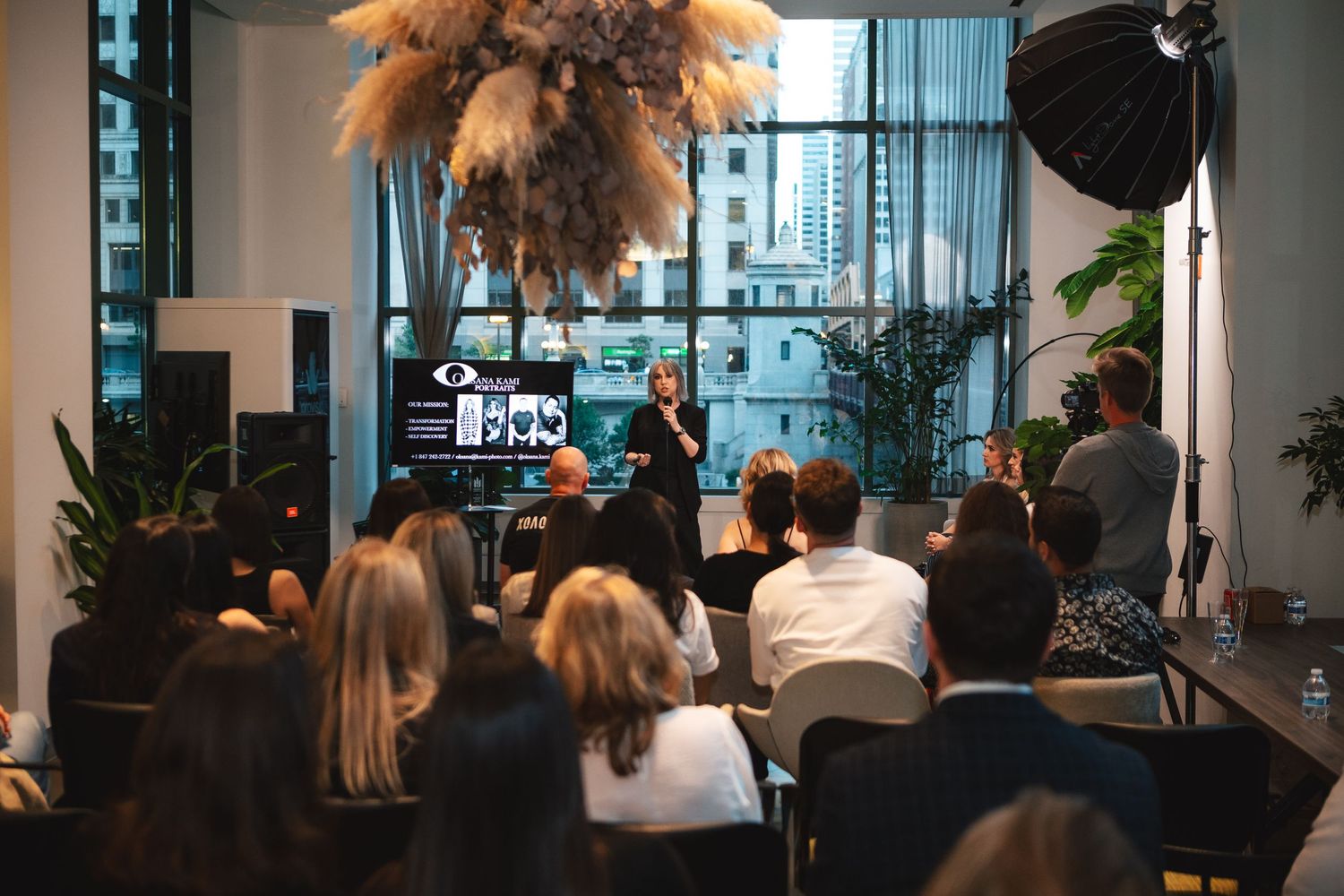 View of audience members attending a professional event with pampas grass decor and city views through windows.