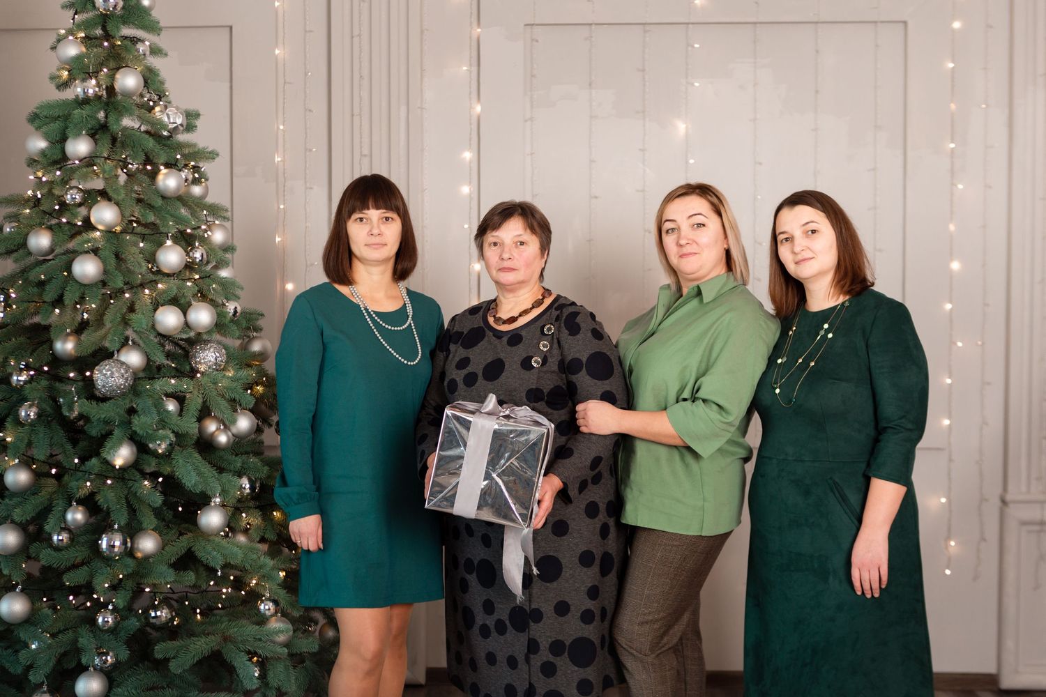Four people in green and black outfits pose with gift by Christmas tree with twinkling lights.