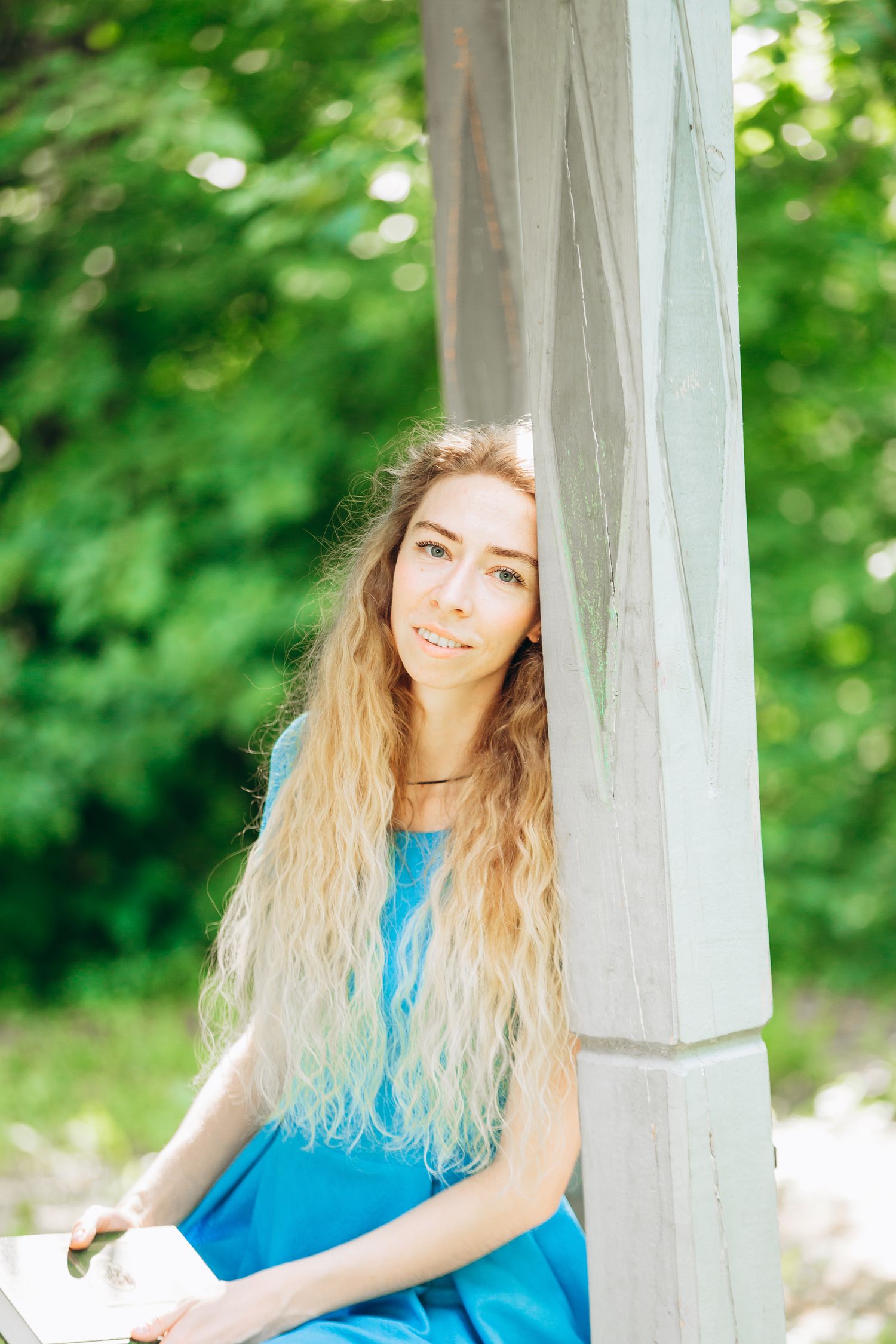Woman with long blonde hair in blue dress sitting against white column outdoors.