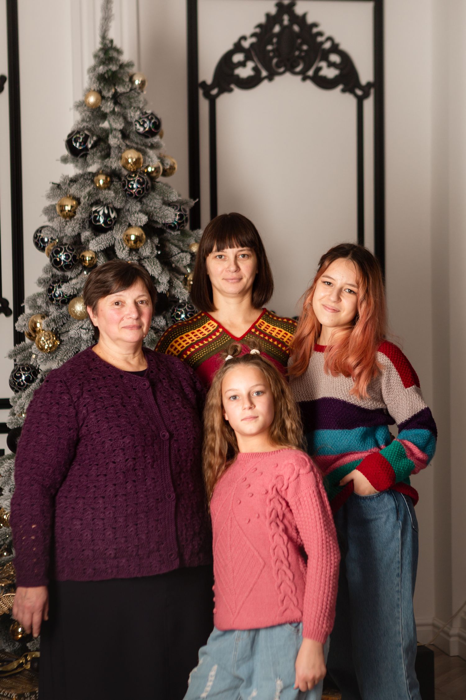 Four generations of women pose by decorated Christmas tree in cozy winter sweaters.