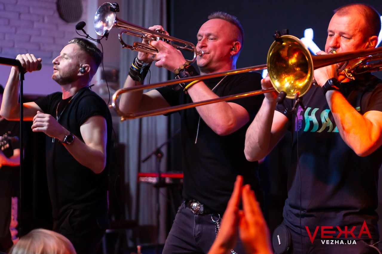 Brass section musicians perform with trombones on stage during a high-energy concert performance.
