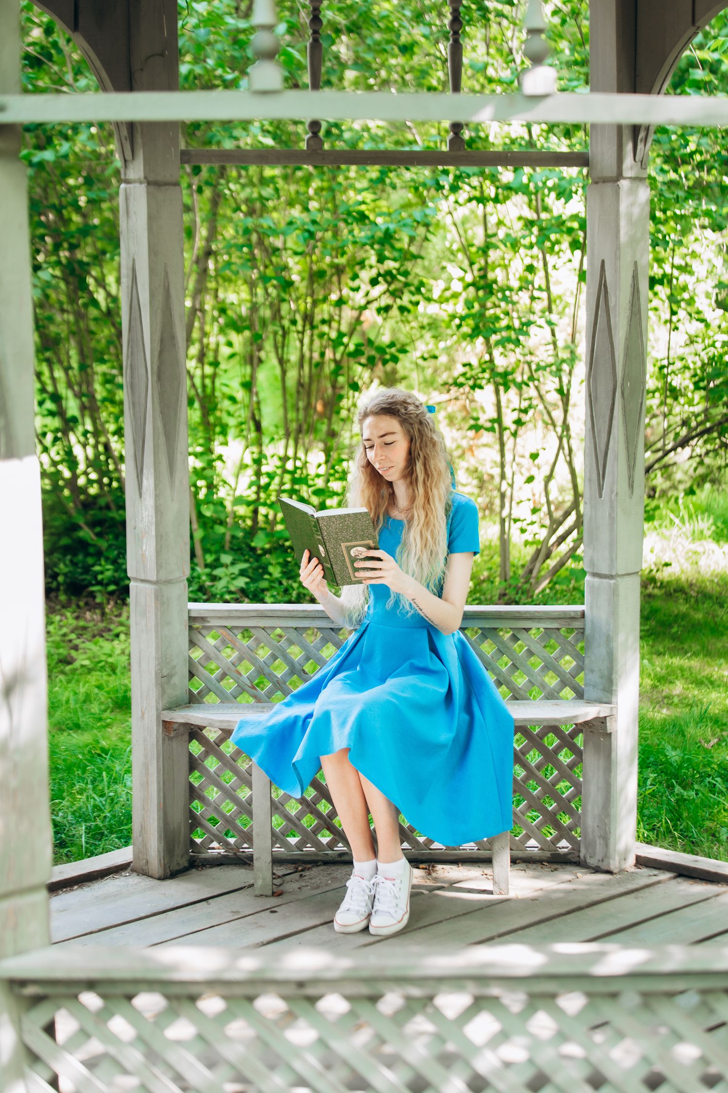 Woman in blue dress reading on wooden bench in garden gazebo with green foliage backdrop.