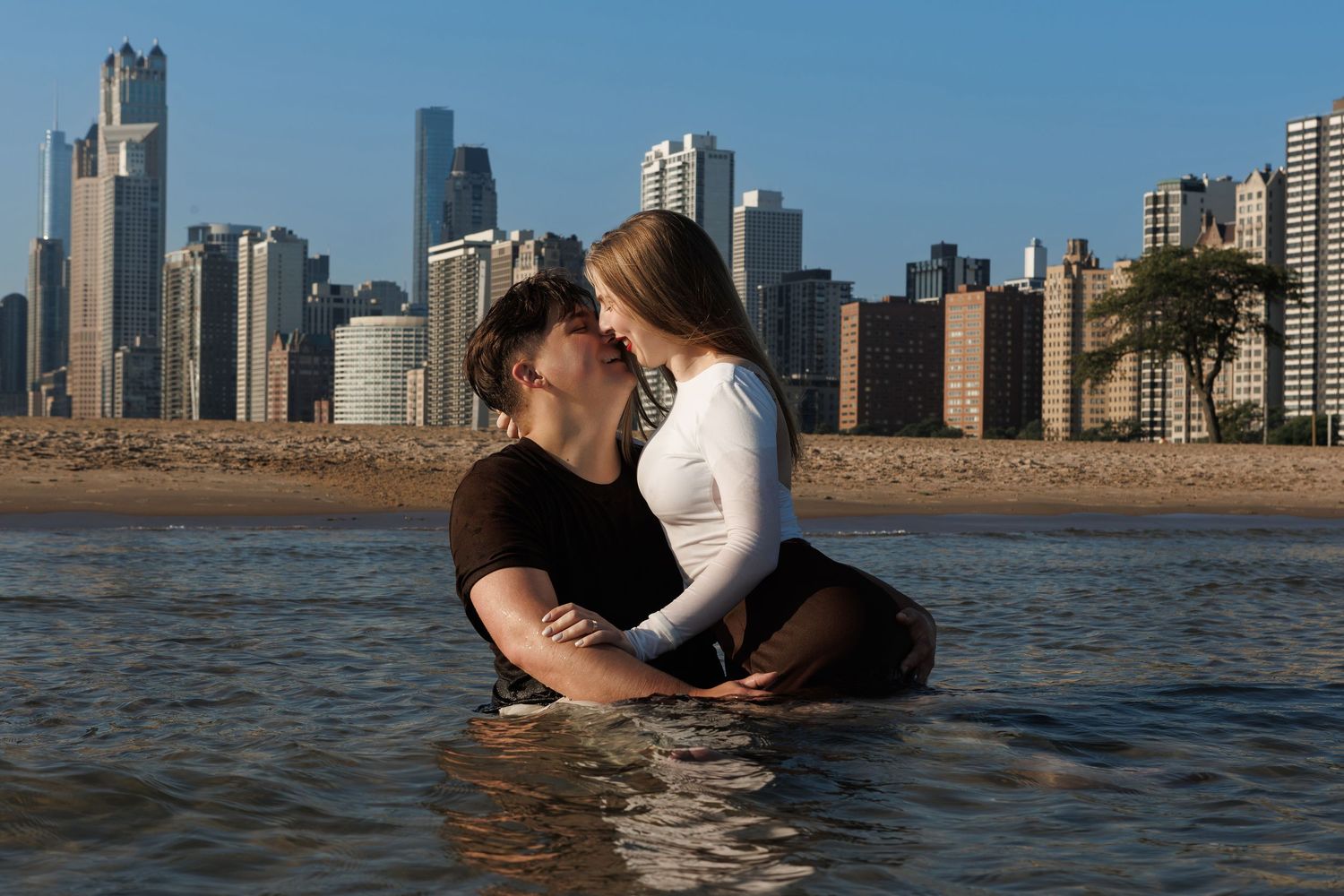 A couple embraces in the water with a city skyline visible in the background during sunset.