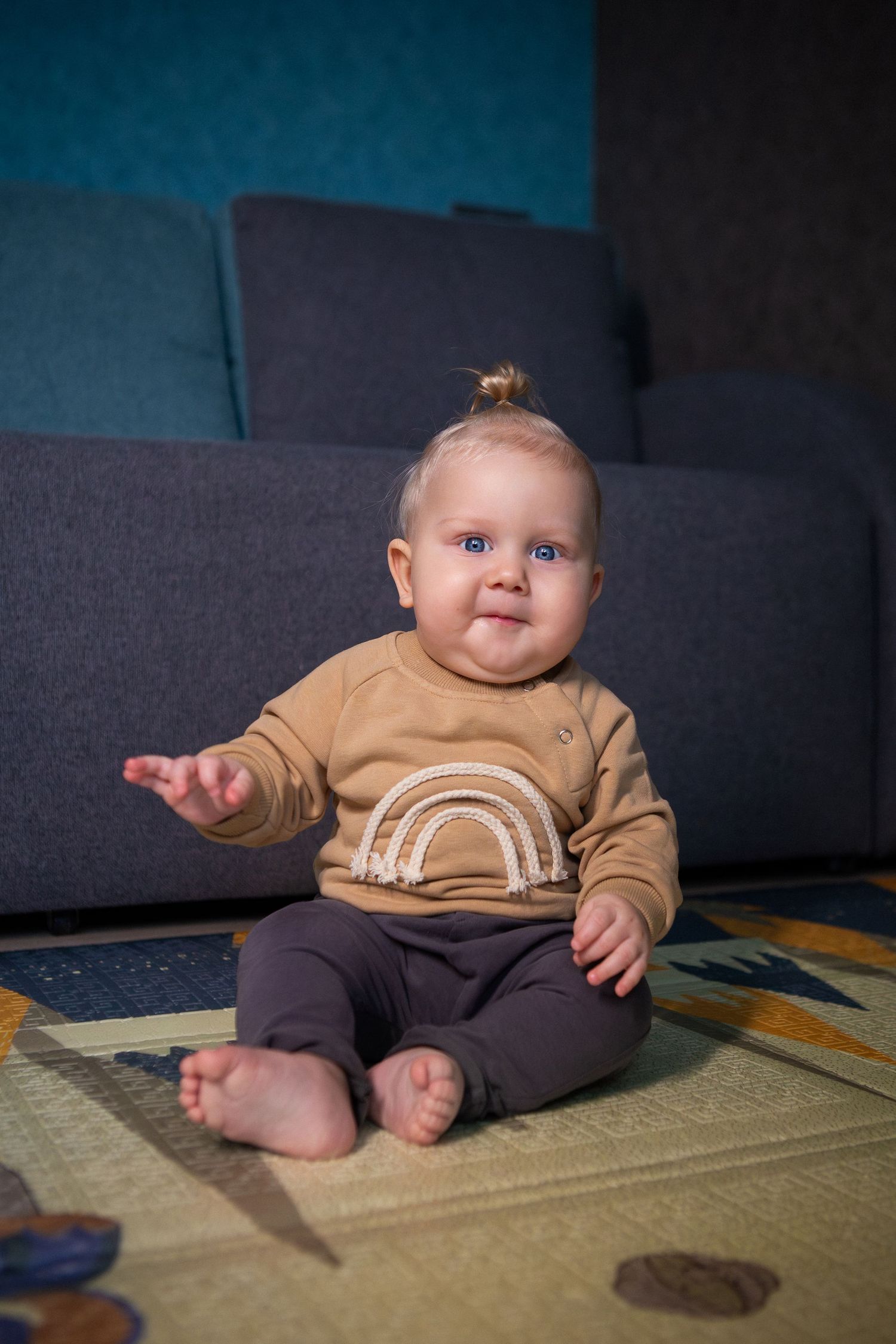 A baby wearing a brown rainbow sweater sits on a patterned rug in front of a dark gray couch.