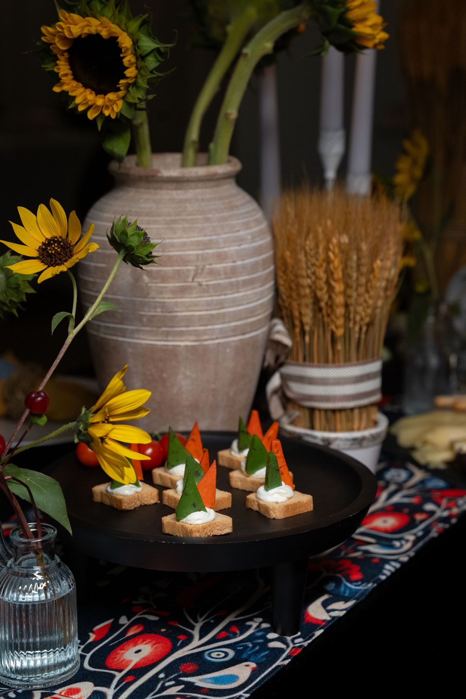 Sunflowers in clay vase with appetizers and wheat stalks on patterned tablecloth.