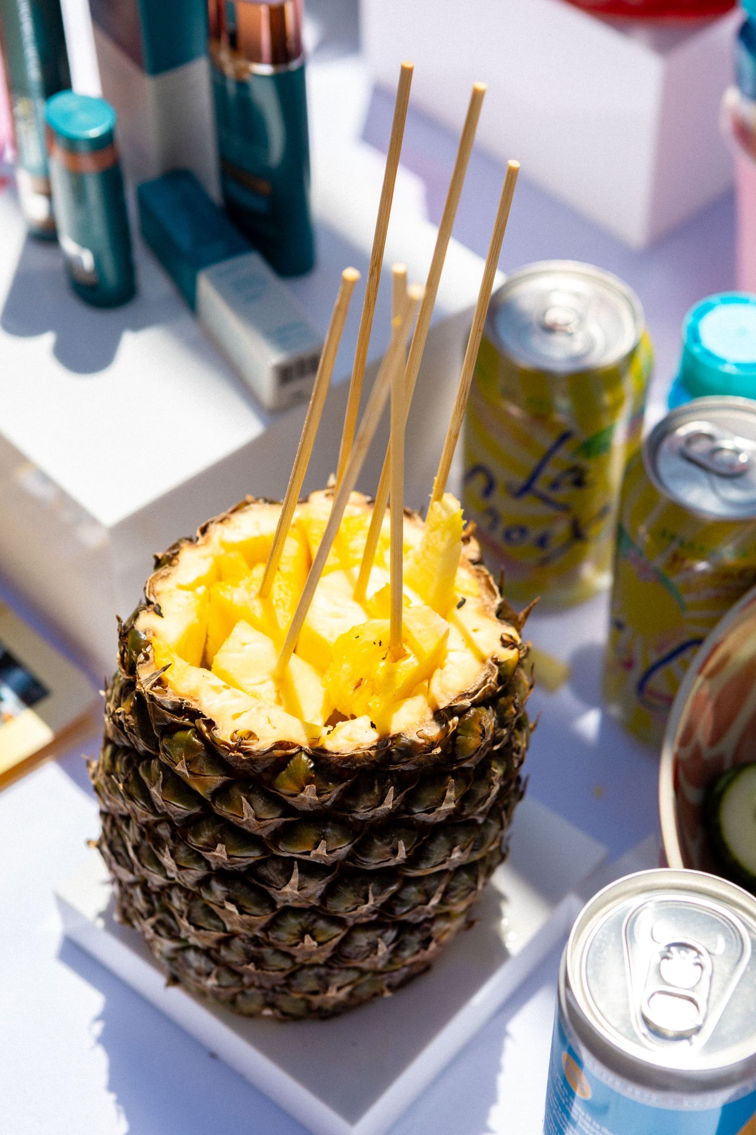 A fresh pineapple bowl filled with diced fruit skewered with bamboo sticks sits next to beverage cans in sunlight.