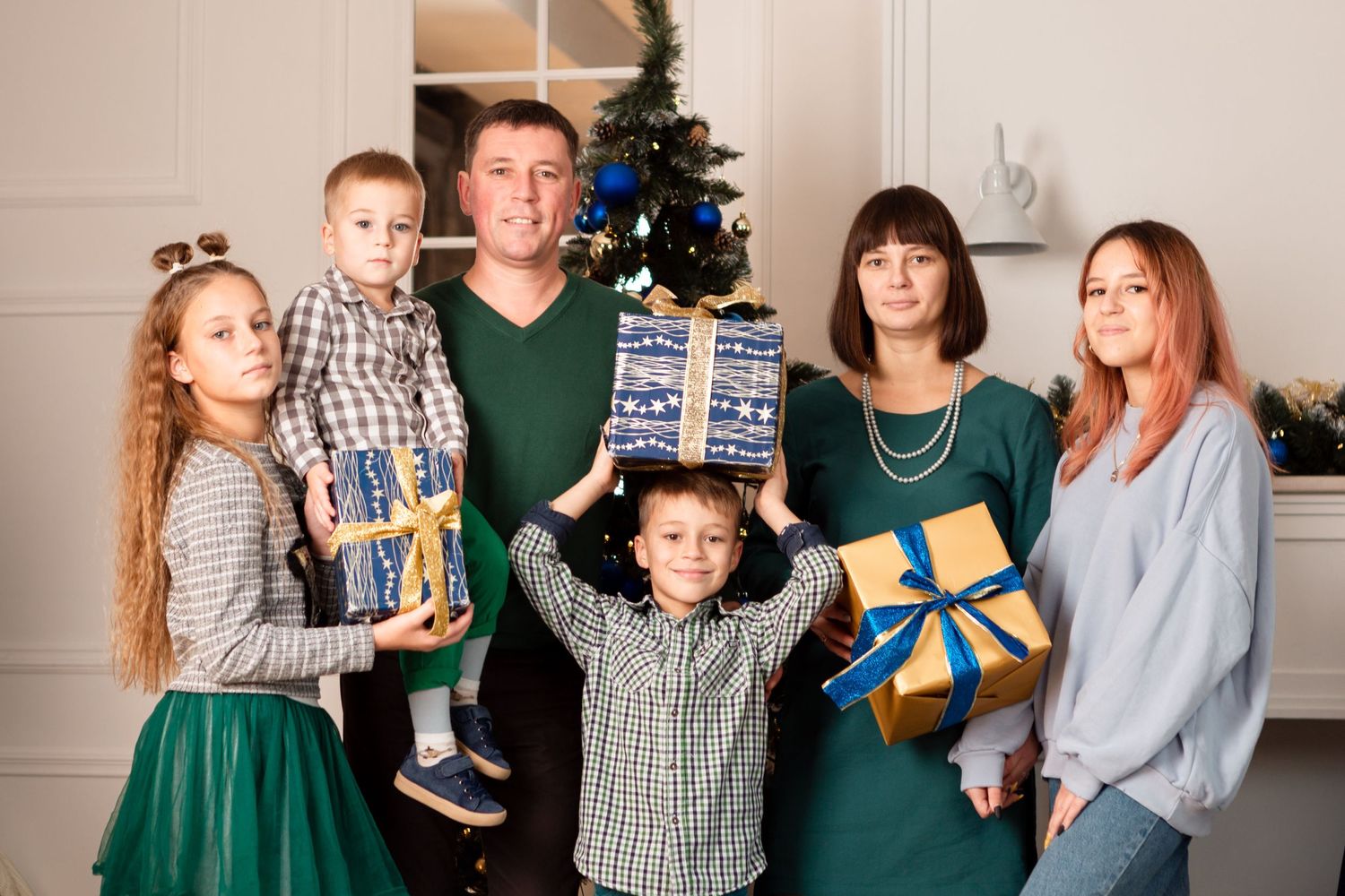 Family group holding wrapped Christmas presents in front of decorated tree.