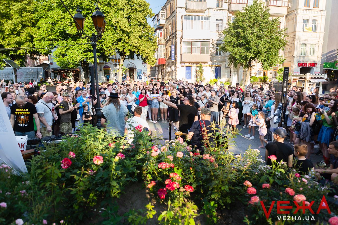Large crowd gathering in urban park with blooming flowers for outdoor concert.